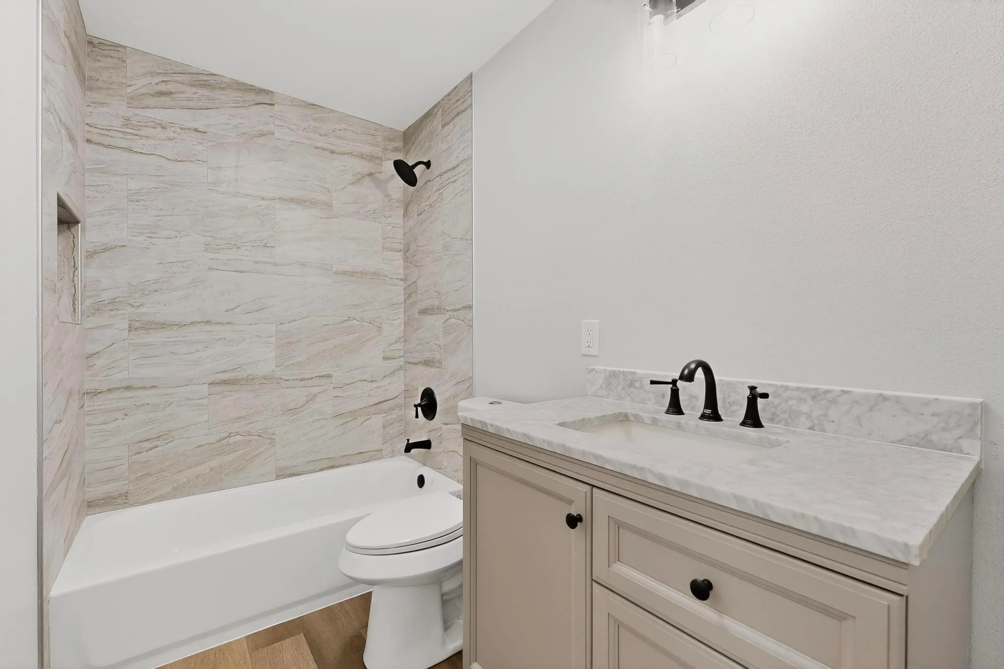Bathtub with tiled surround and built-in shower niche, matte black shower fixtures, vanity with light-colored countertop, matte black sink faucet, and wood-finish​​‌​​​​‌​​‌‌​‌‌​​​‌‌​‌​‌​‌​​​‌​​ flooring