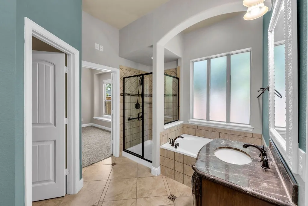 Tiled flooring with accent details, walk-in shower with glass enclosure, separate soaking tub, vanity with stone countertop and integrated sink, and an arched entryway
