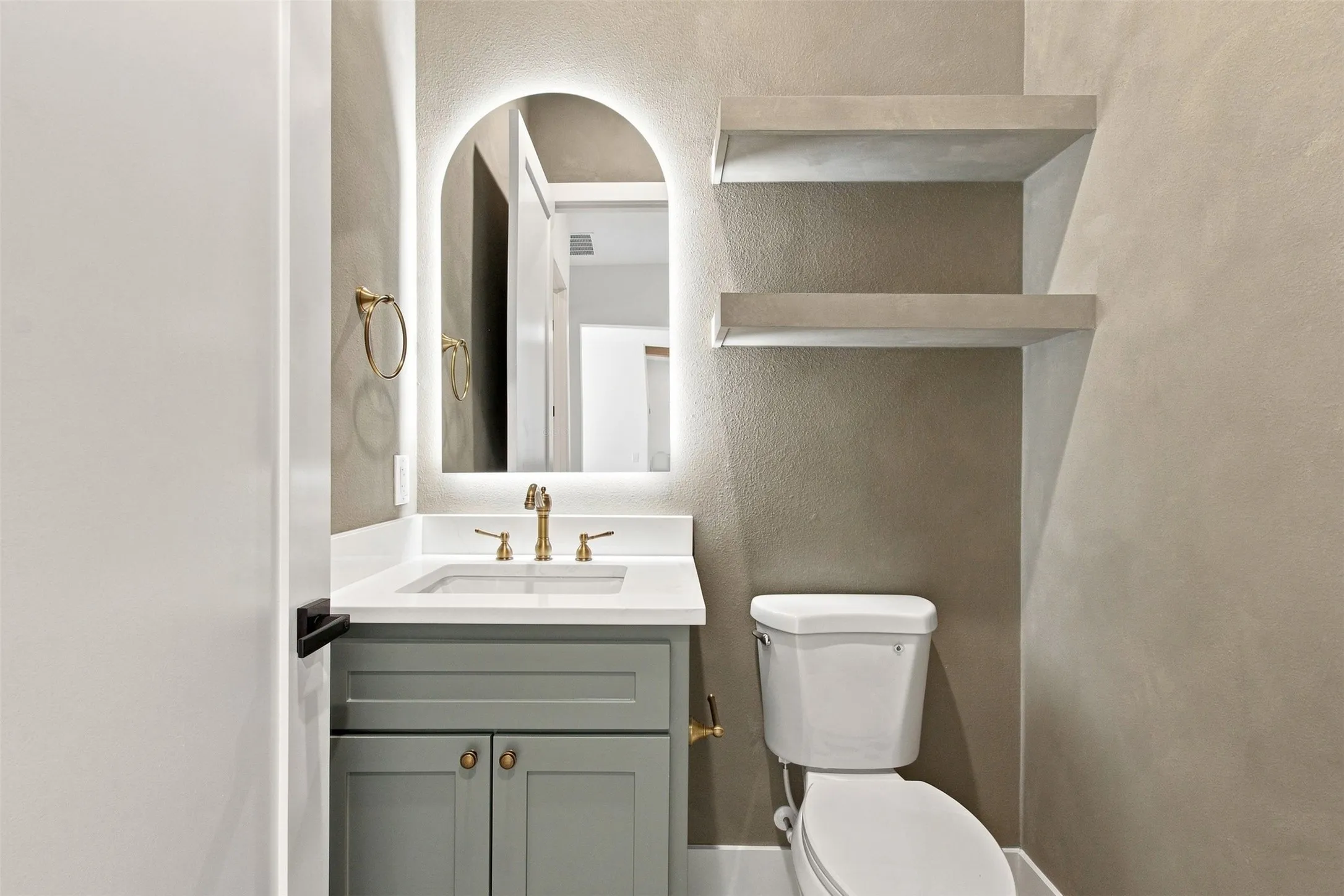Bathroom featuring a vanity with a white countertop, an arched backlit mirror, gold-finish hardware, and built-in floating shelves