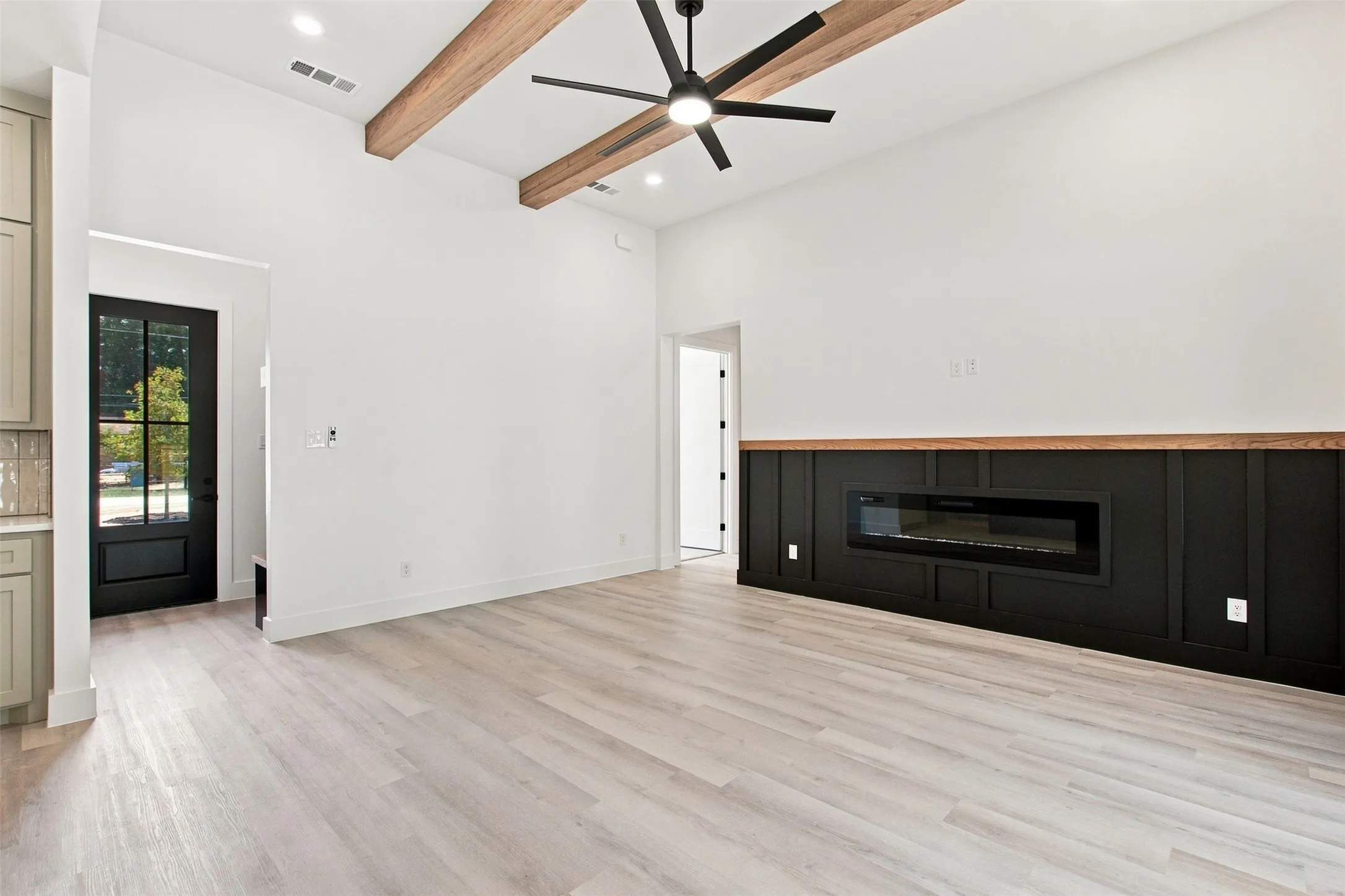 Vaulted ceilings with exposed wood beams, plank wood-finish flooring, linear electric fireplace, paneled accent wall, and a ceiling fan