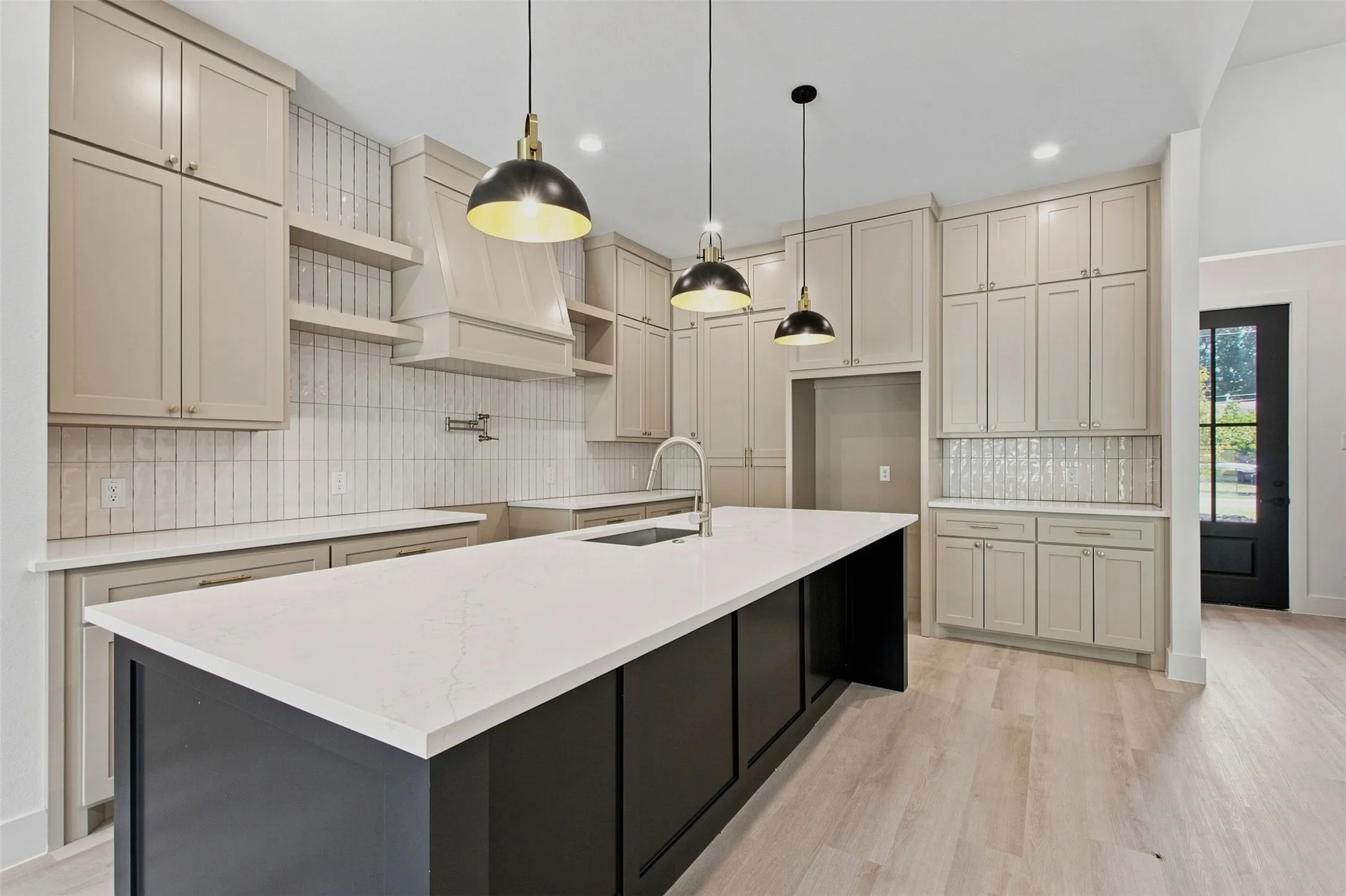 Spacious kitchen featuring a large island with a light-toned countertop, extensive light-toned cabinetry, light wood-finish flooring, a decorative hood, and a full-height tile backsplash