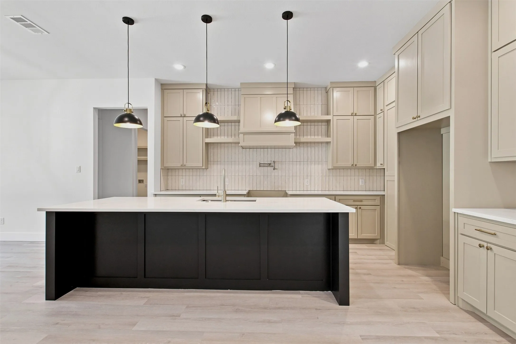 Spacious kitchen featuring a large black island with white countertop, light wood-finish flooring, cream-colored cabinetry, and a full-height tile backsplash