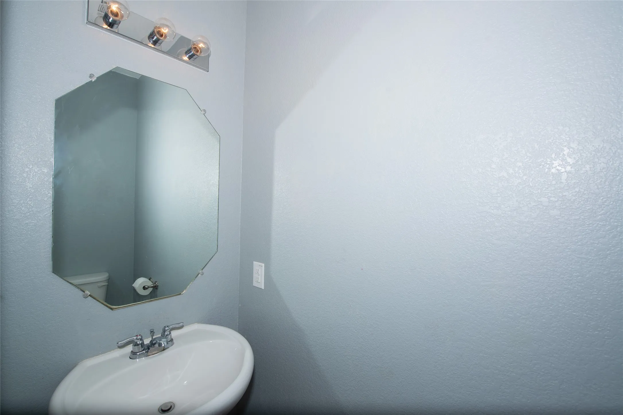 Pedestal sink with chrome-finish faucet, octagonal wall mirror, and three-light vanity fixture