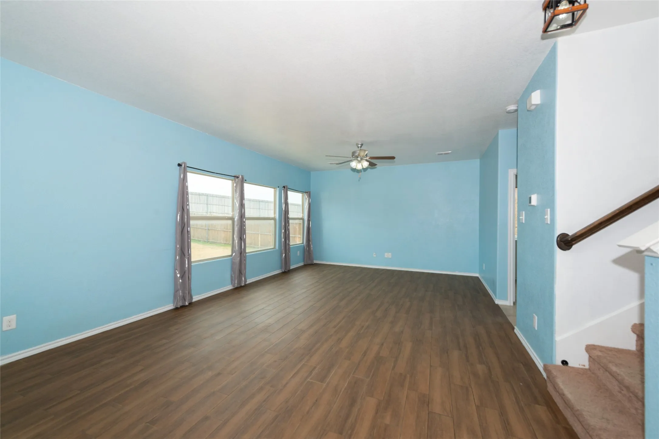 Spacious room featuring wood-finish flooring, three large windows, a ceiling fan, and a staircase with a dark wood handrail