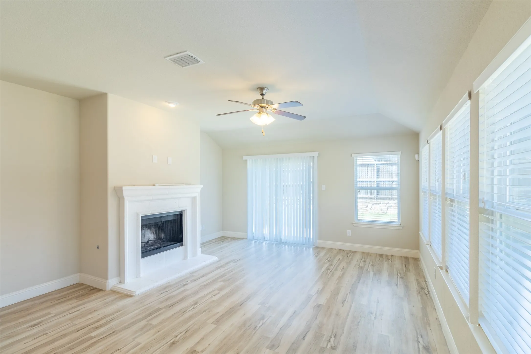 Spacious living area featuring wood-finish flooring, a white mantel fireplace, vaulted ceiling, and multiple windows with blinds