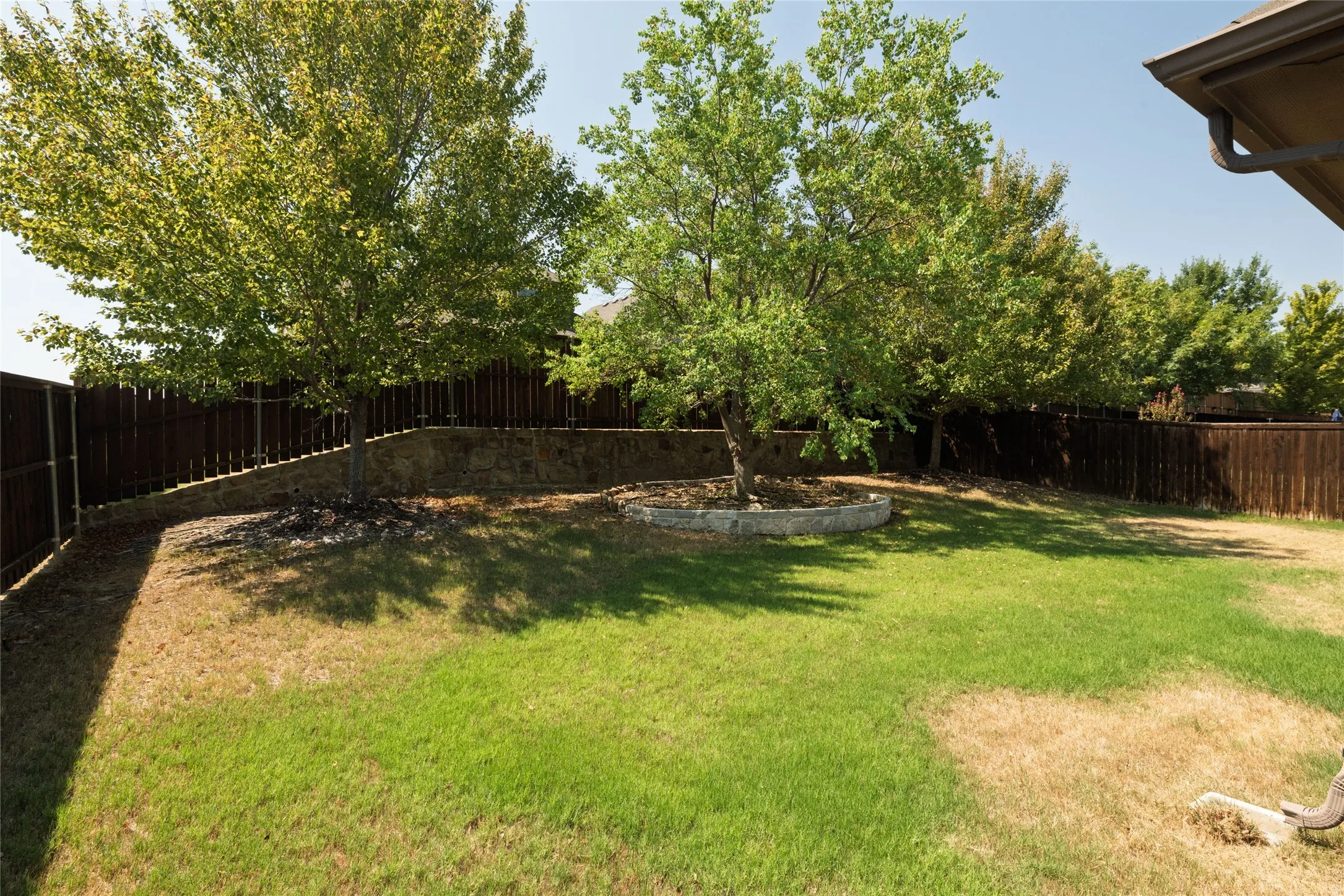 Expansive rear yard featuring green lawn, mature trees, wood privacy fencing, and a stone retaining wall