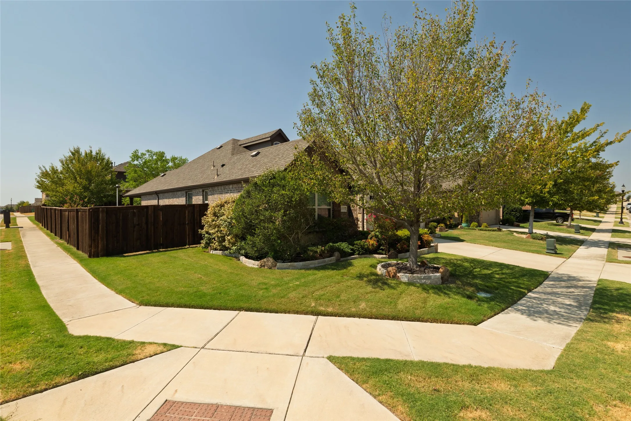 Brick exterior with a dark shingle roof, mature trees, landscaped shrubbery, and a wood privacy fence