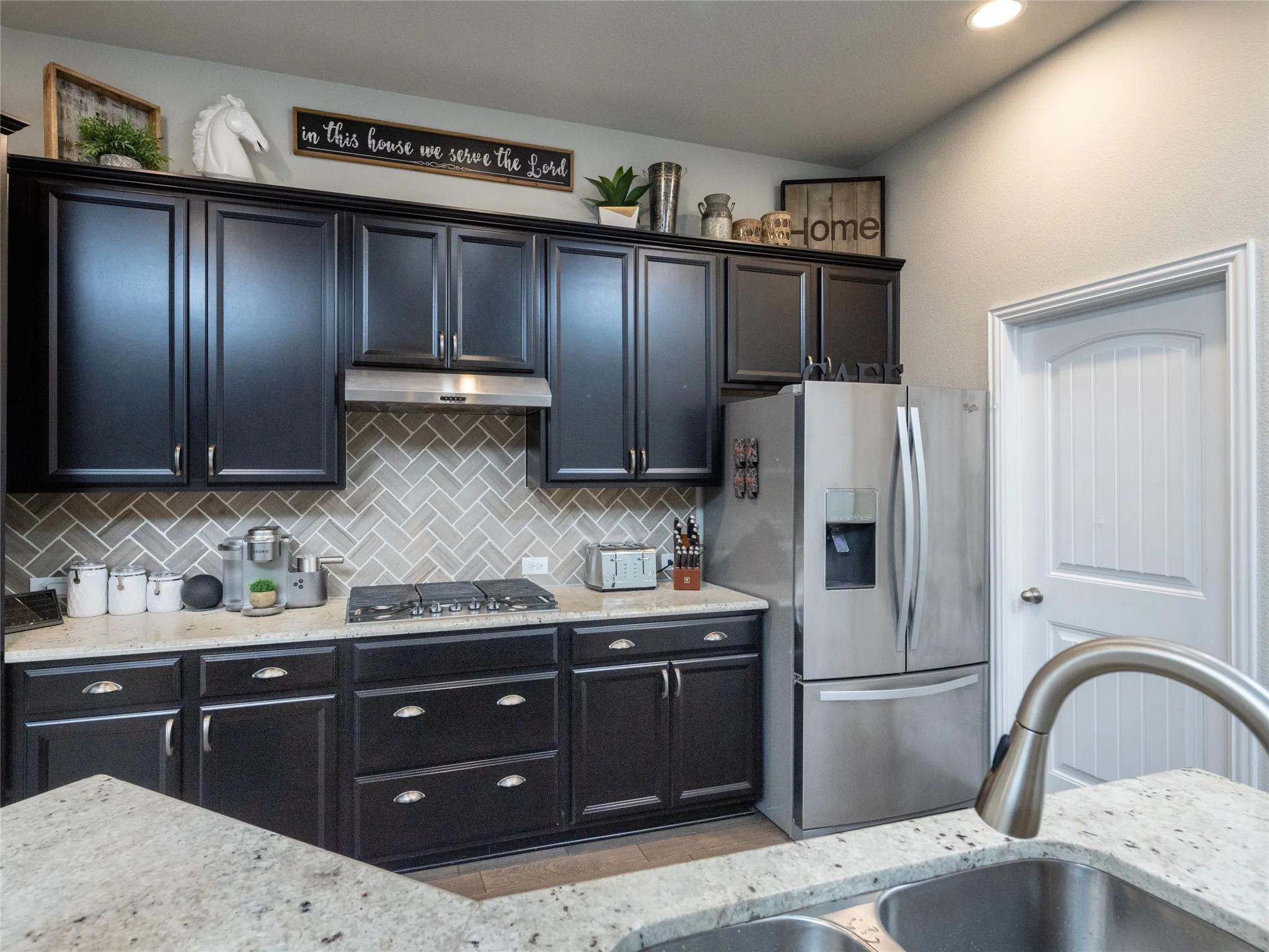 Dark cabinetry with crown molding, stainless steel appliances, herringbone tile backsplash, and light-toned countertops