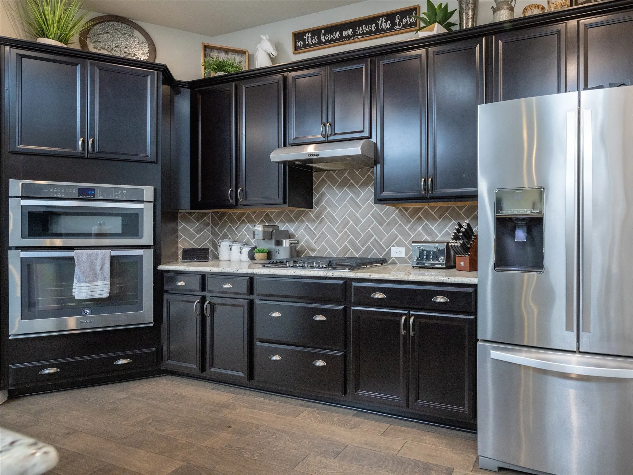 Black cabinetry with silver hardware, light-toned countertops, herringbone pattern tile backsplash, stainless steel double wall oven, and wood-finish flooring