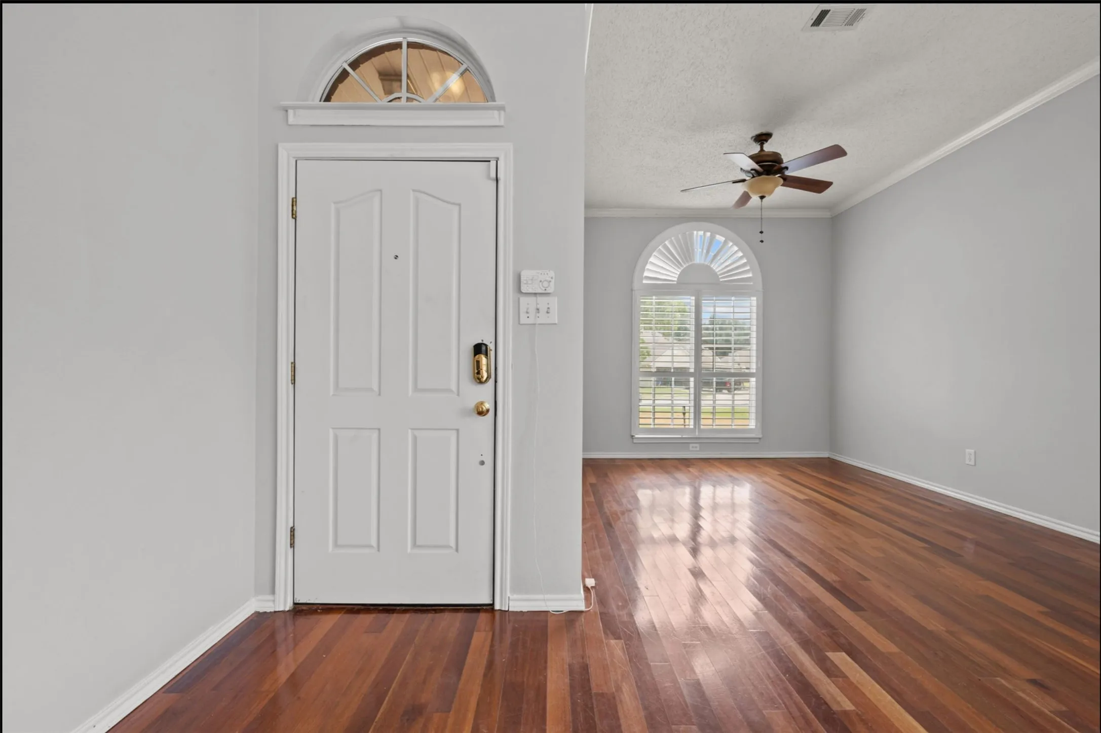 Wood flooring, white paneled door with an arched transom, arched window with plantation shutters, ceiling fan, and crown molding