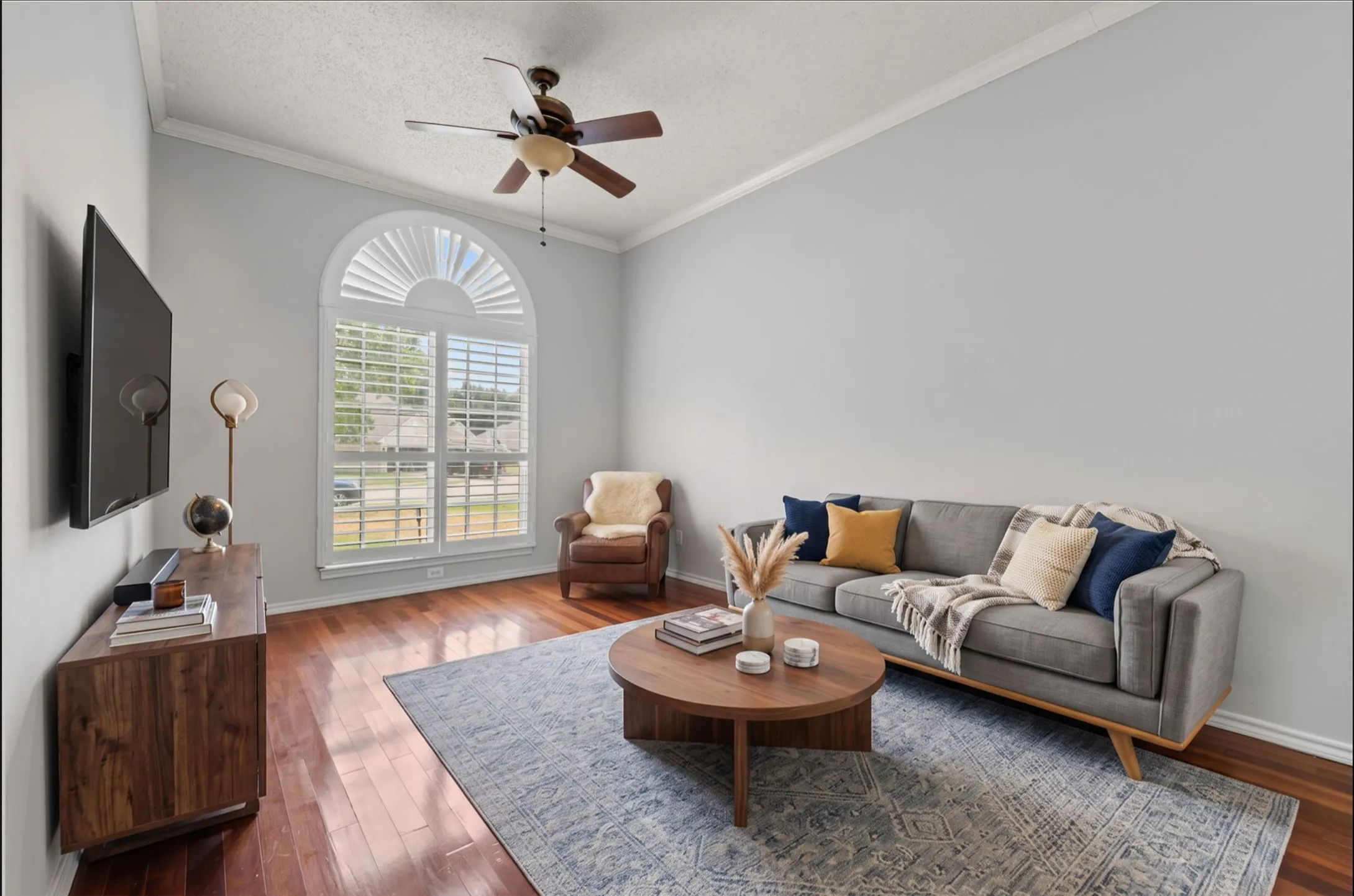 Arched window with louvered plantation shutters crown molding, wood flooring, ceiling fan with light.