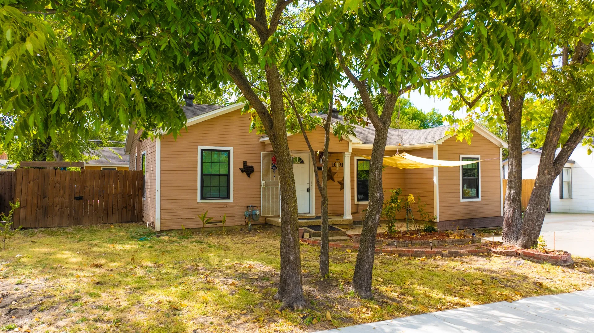 Siding exterior, multiple mature trees, front porch with columns, white front door, and a partial privacy fence