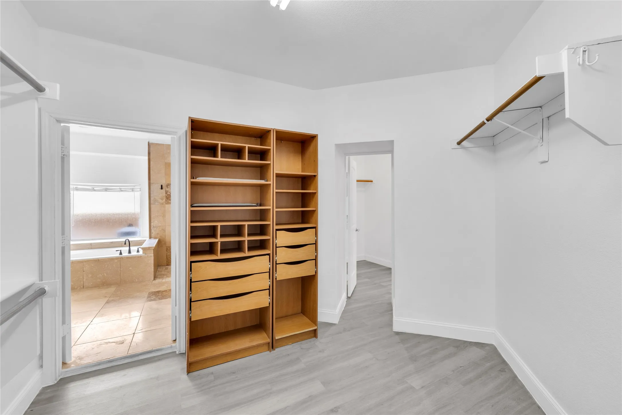 Integrated closet organization system, additional shelving, light wood-finish flooring, and a stone-tiled bathroom with a built-in tub