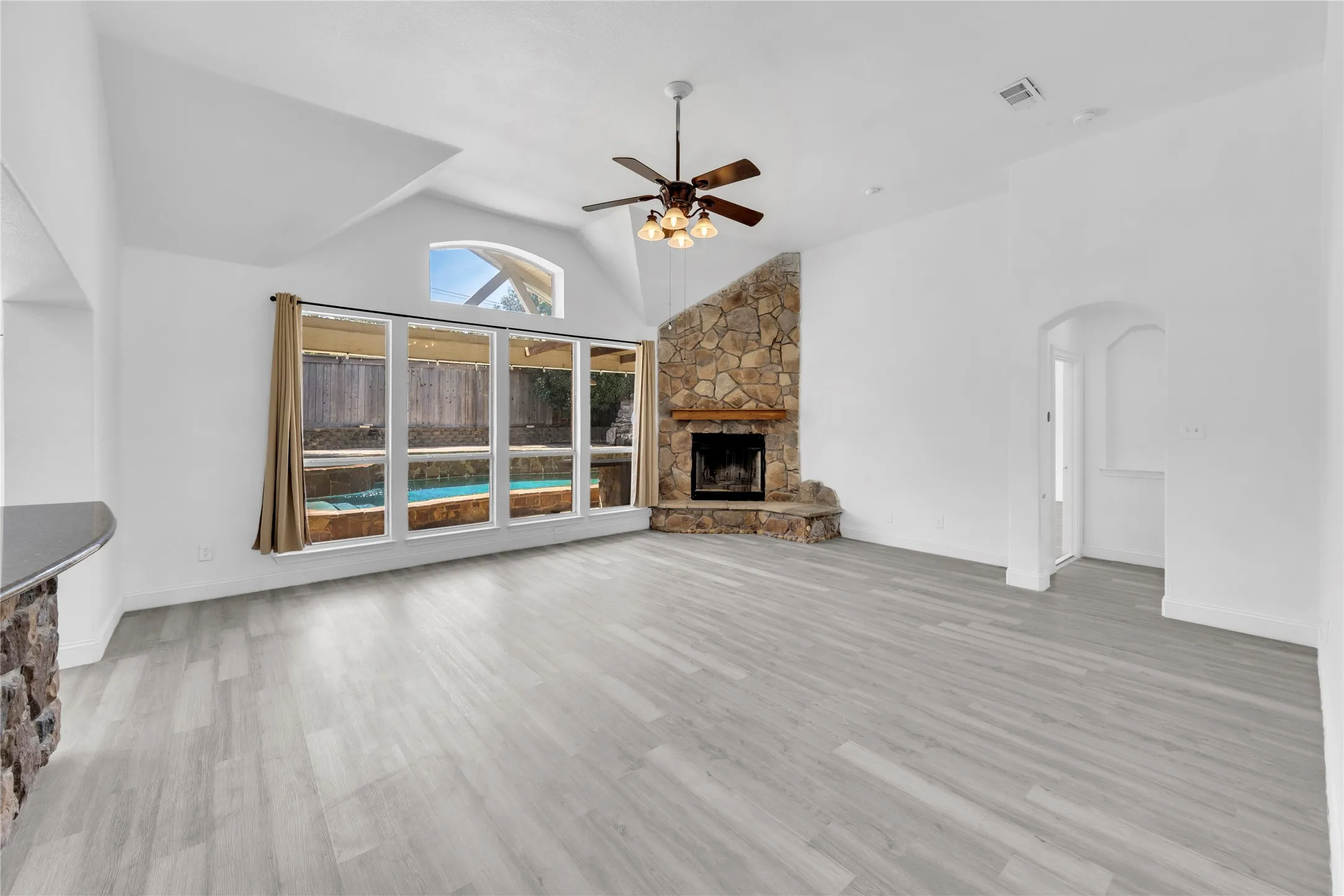Vaulted ceilings with a ceiling fan, stone-clad corner fireplace, wood-finish flooring, and a window wall overlooking a pool