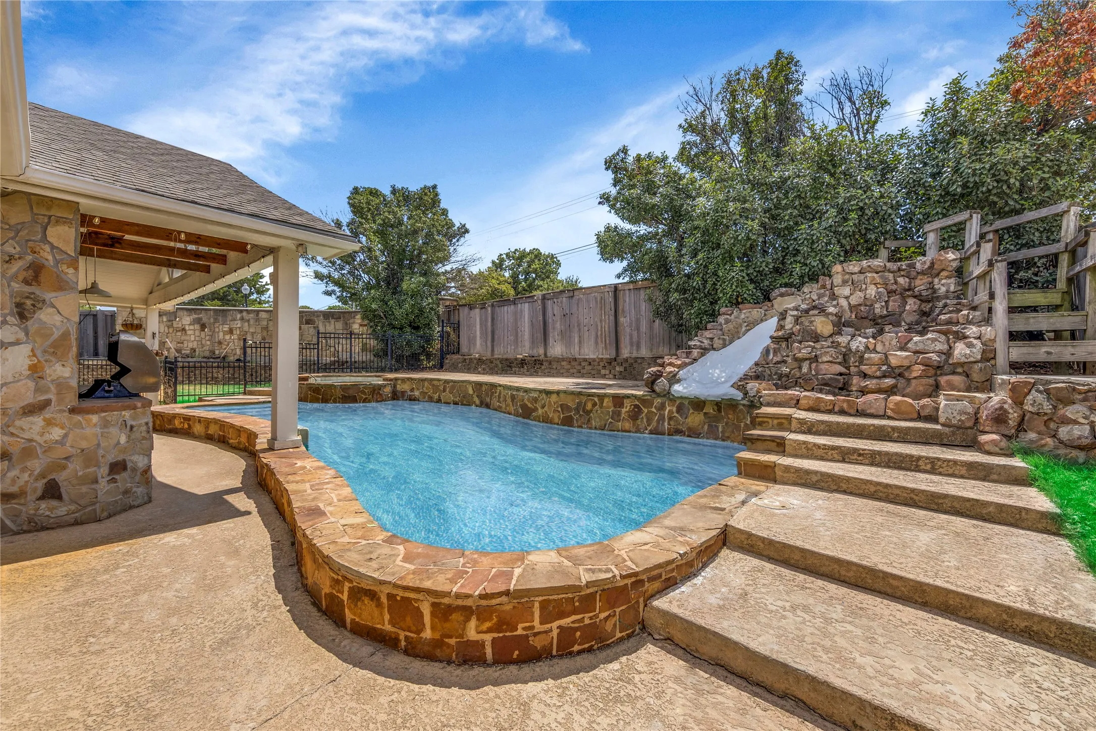 Outdoor oasis featuring a freeform swimming pool, stone-clad water slide, concrete patio, and a covered patio with stone pillar accents