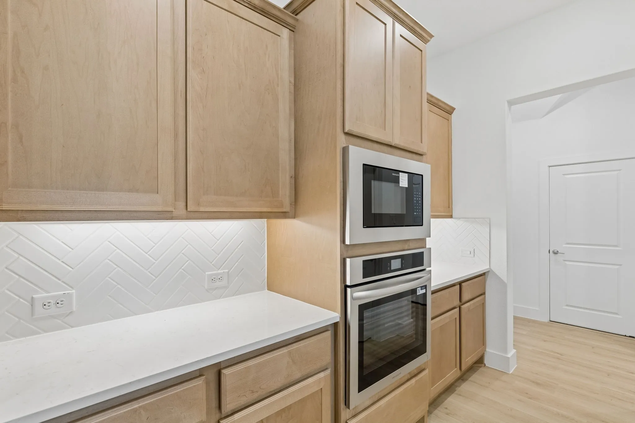 Light wood-finish cabinetry with crown molding, stainless steel built-in microwave and oven, white countertops, and white herringbone tile backsplash