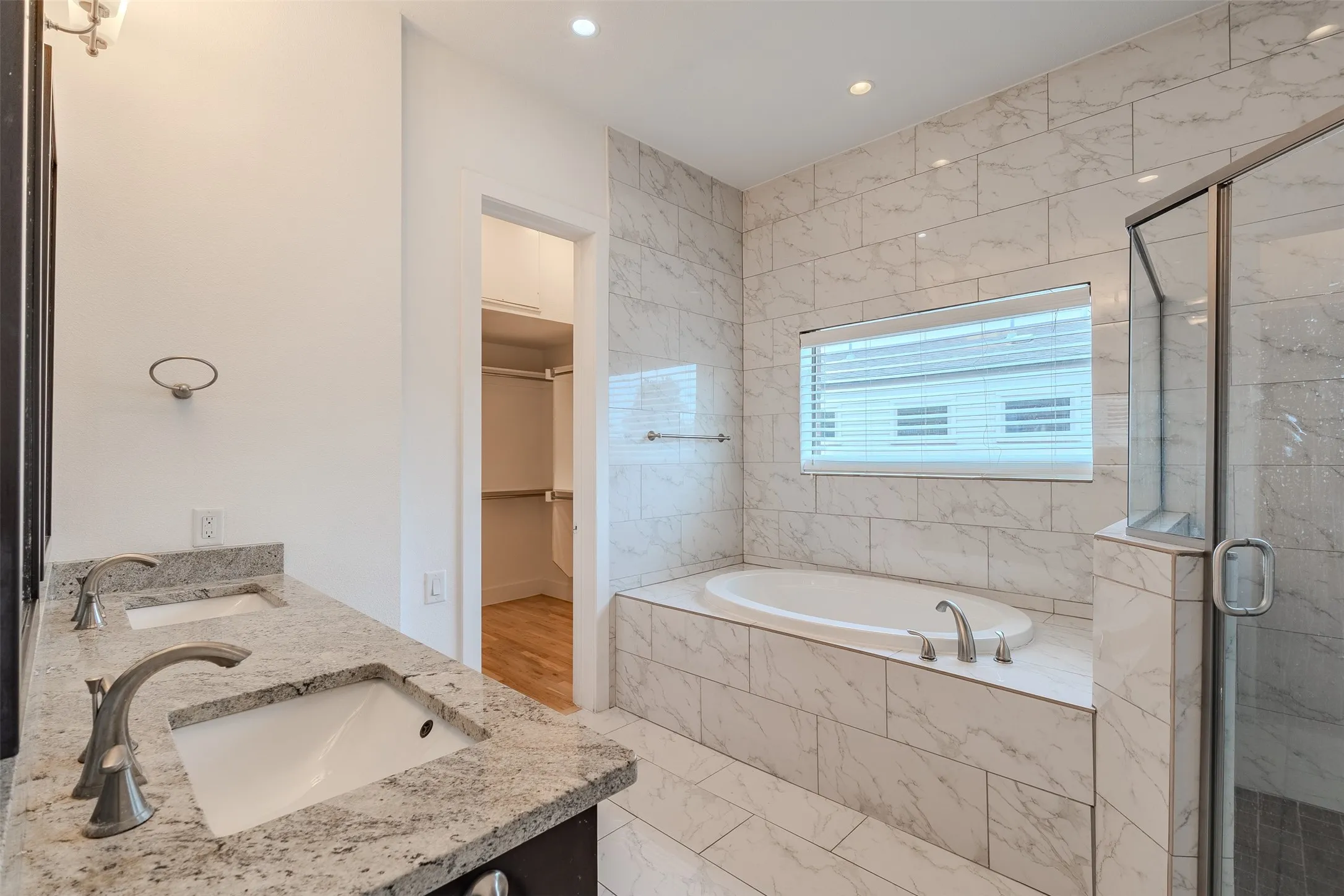 Dual vanity with undermount sinks and light-toned stone countertops, oversized soaking tub with tile surround, glass-enclosed shower, and tiled flooring