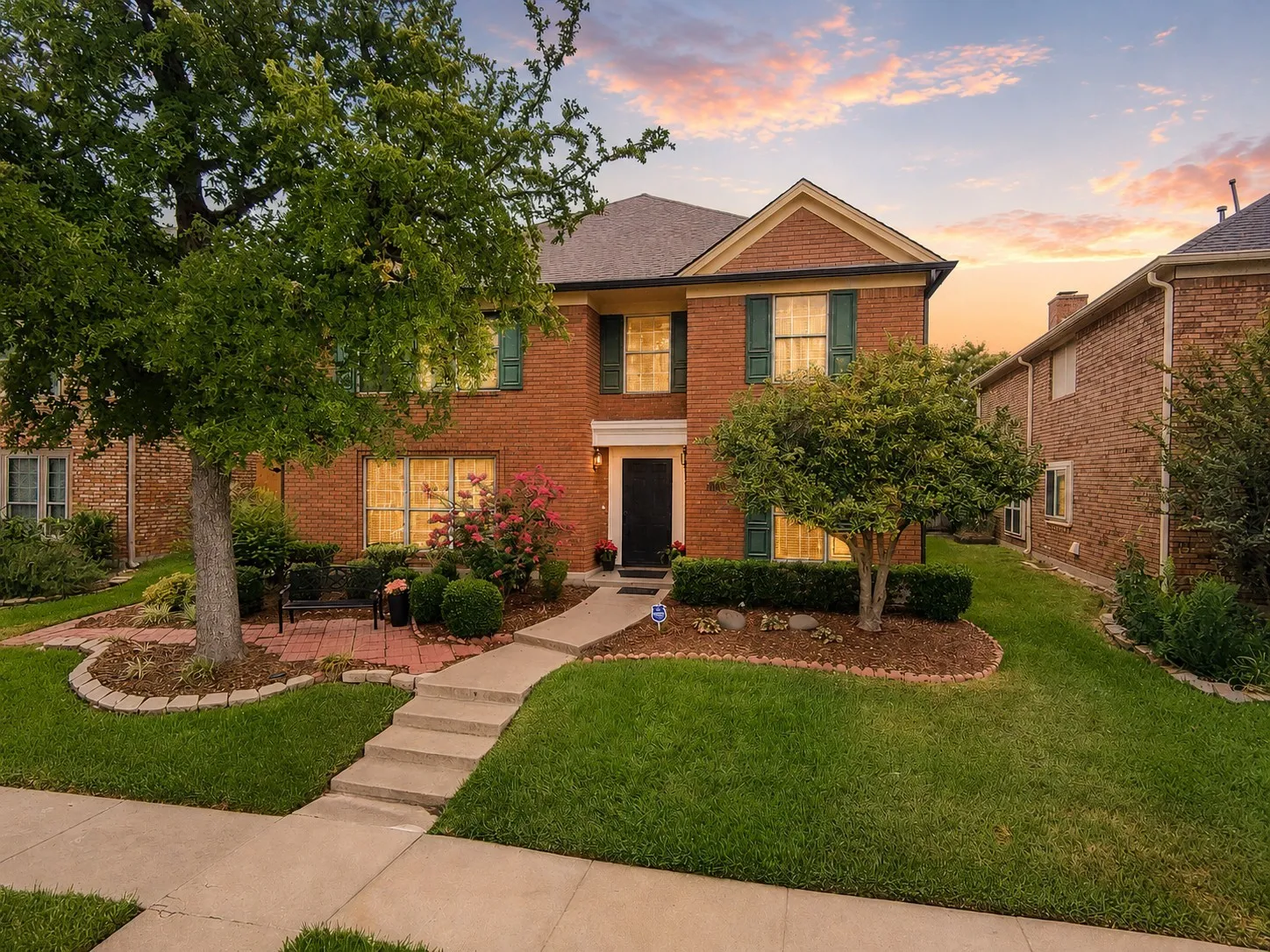 Two-story brick exterior with green shutters, a concrete walkway, mature trees, and landscaping with shrubs and flowering plants