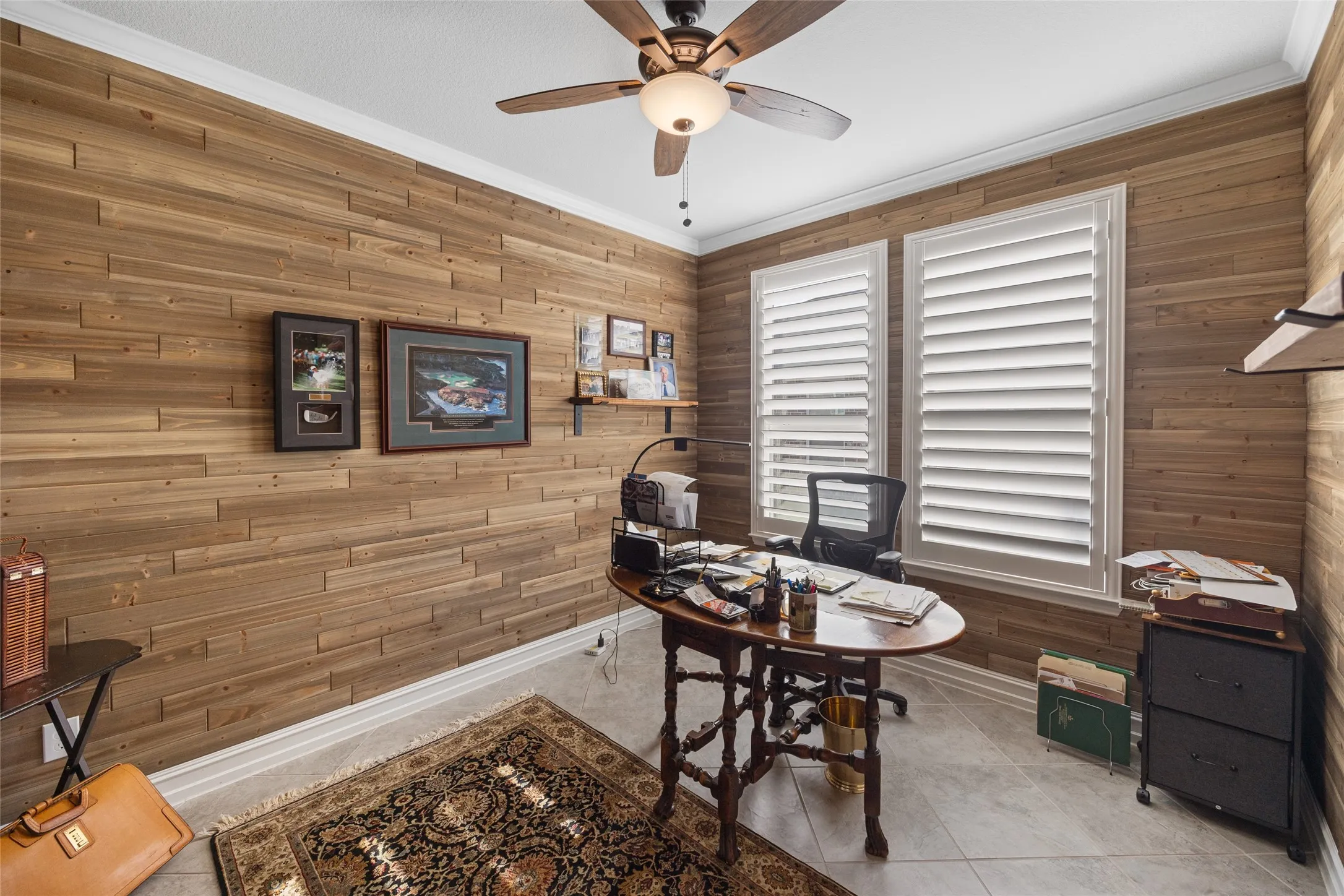 Wood-paneled walls, ceiling fan, crown molding, white window shutters, and tiled flooring