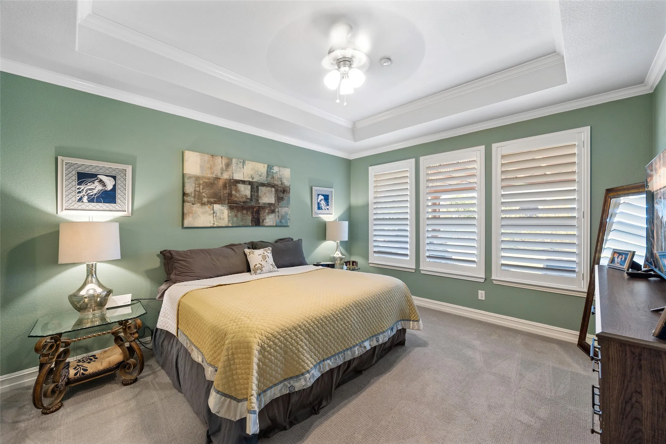 Recessed ceiling with layered crown molding, ceiling fan light fixture, three windows with plantation shutters, and light-toned carpet flooring