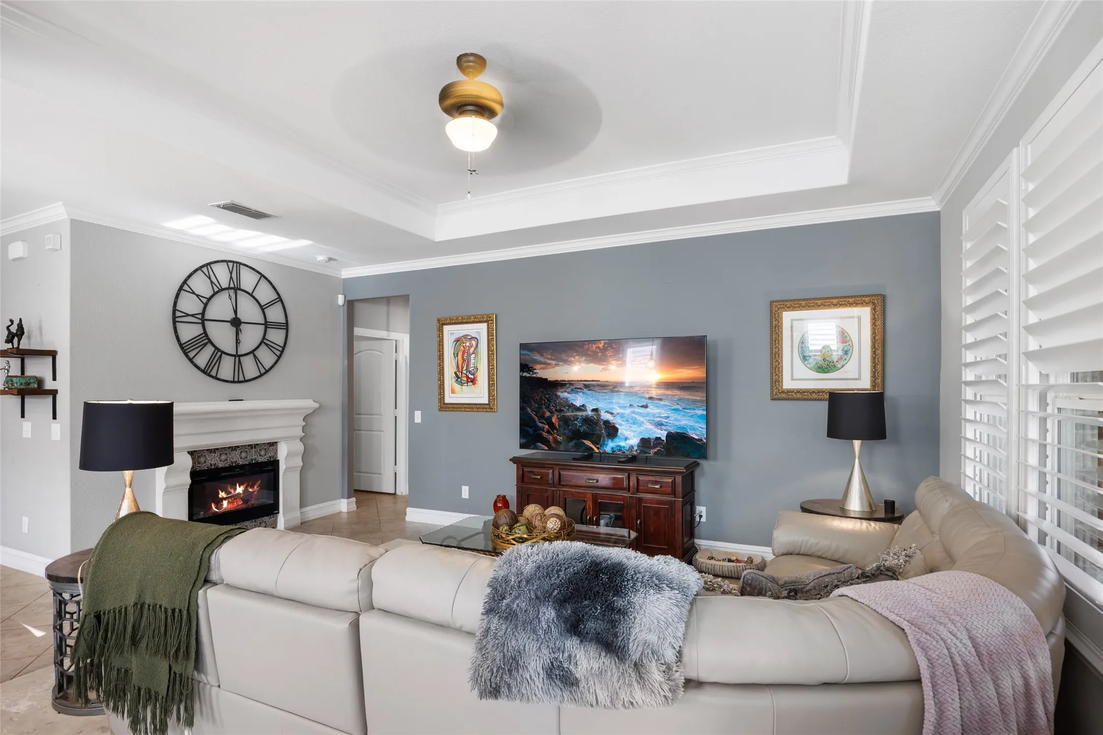 Tray ceiling with a ceiling fan, crown molding, fireplace with a decorative mantel, and plantation shutters