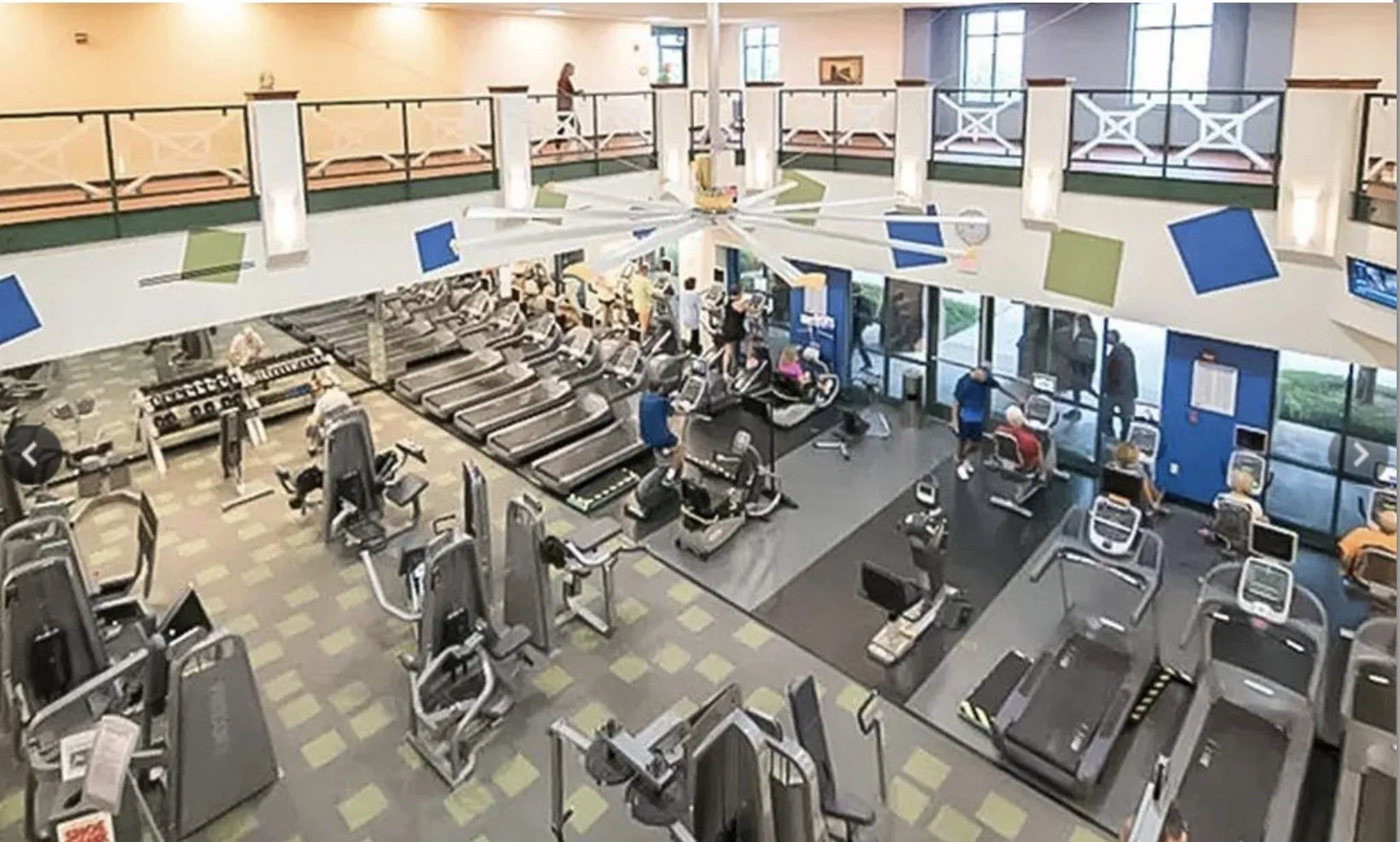 Two-story fitness center with extensive exercise equipment, high ceilings, large windows, and an upper-level walkway with decorative railings