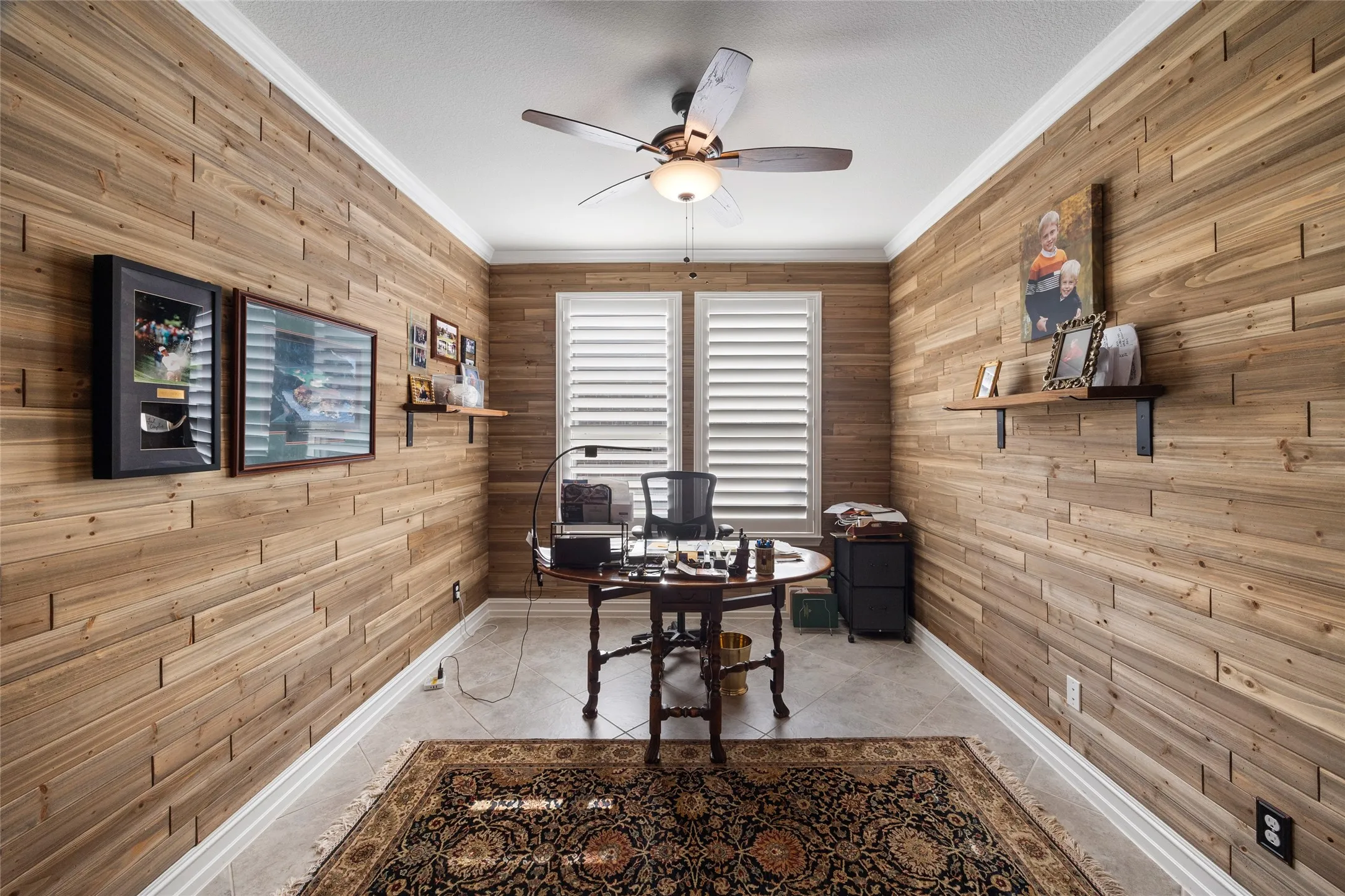 Wood-panel accent walls, ceiling fan with integrated lighting, window with white plantation shutters, white crown molding, and tile flooring