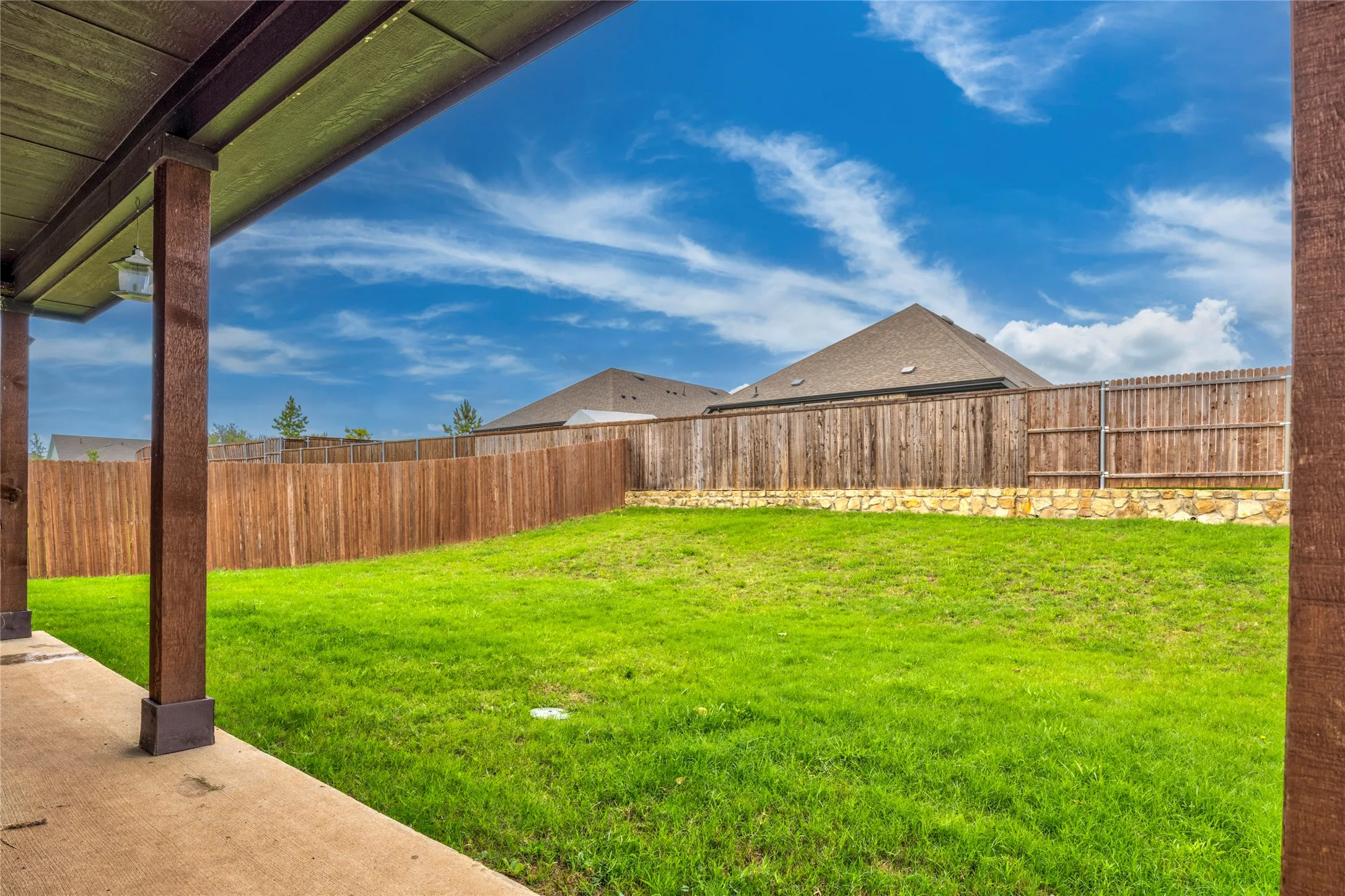 Expansive backyard featuring a lush lawn, privacy fencing with stone base, and covered patio with wood-finish posts