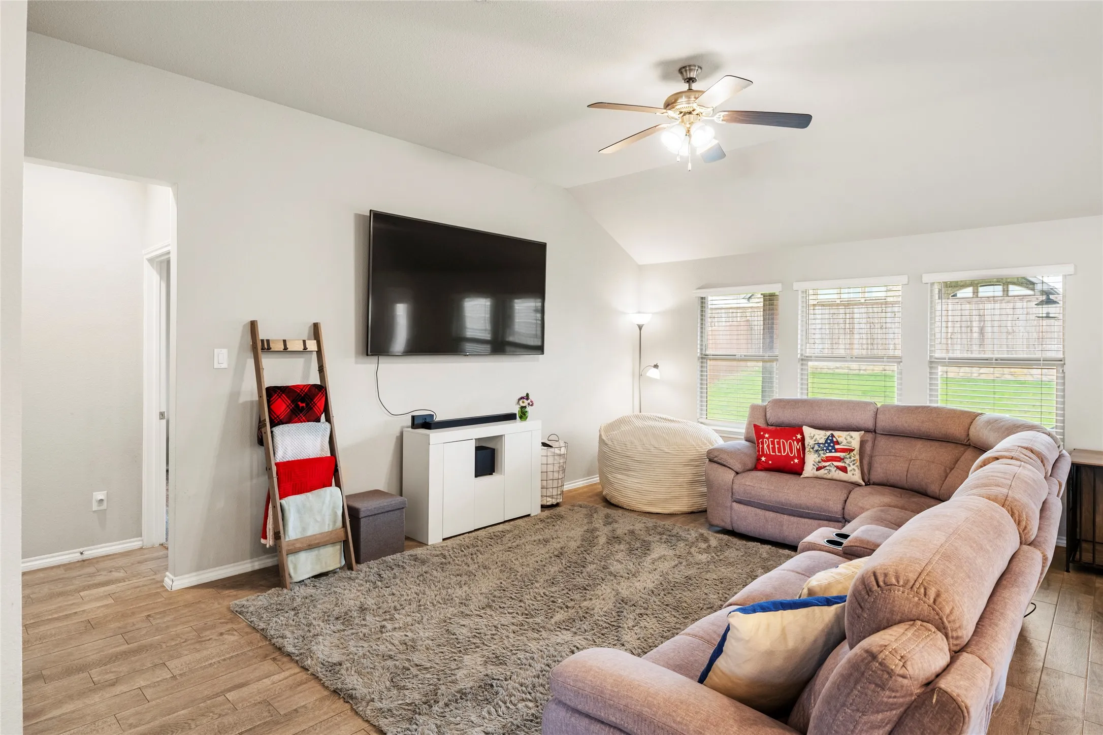 Vaulted ceiling with a ceiling fan, three windows with blinds, looking onto the good sized back yard, wood-like tile flooring, and light wall paint....