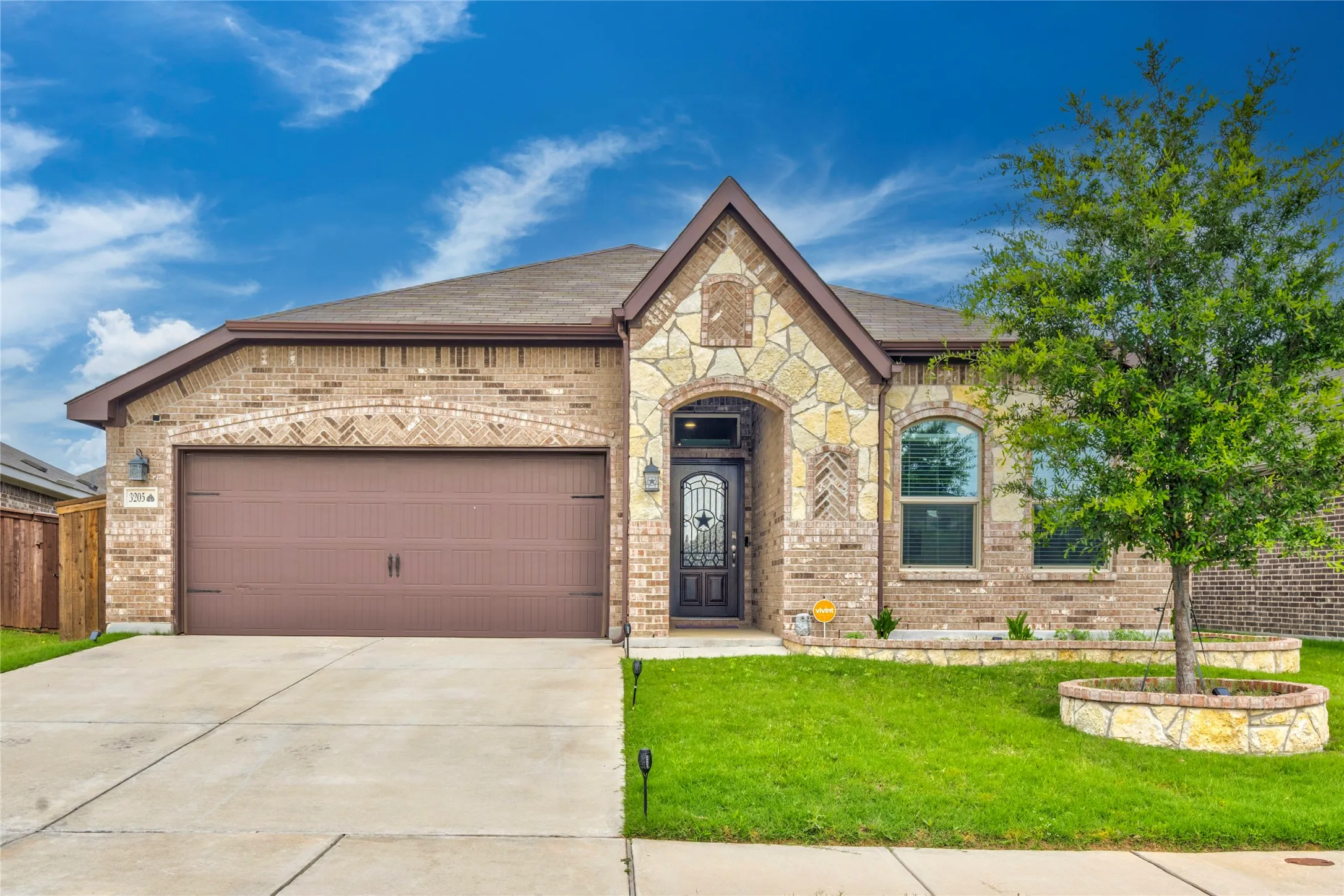 Brick exterior with stone accents, arched entry, decorative front door, attached garage, and a landscaped front yard with a tree