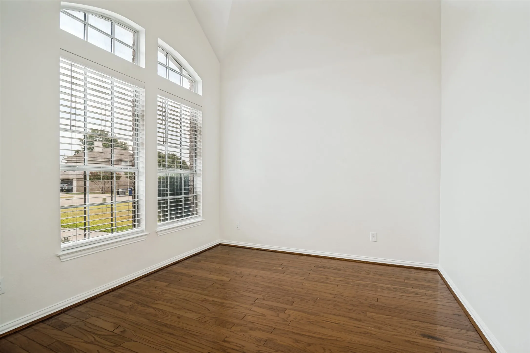 Spacious front living room featuring vaulted ceilings, wood-finish flooring, white walls, and large windows with arched transoms and blinds. Could be used for office space.