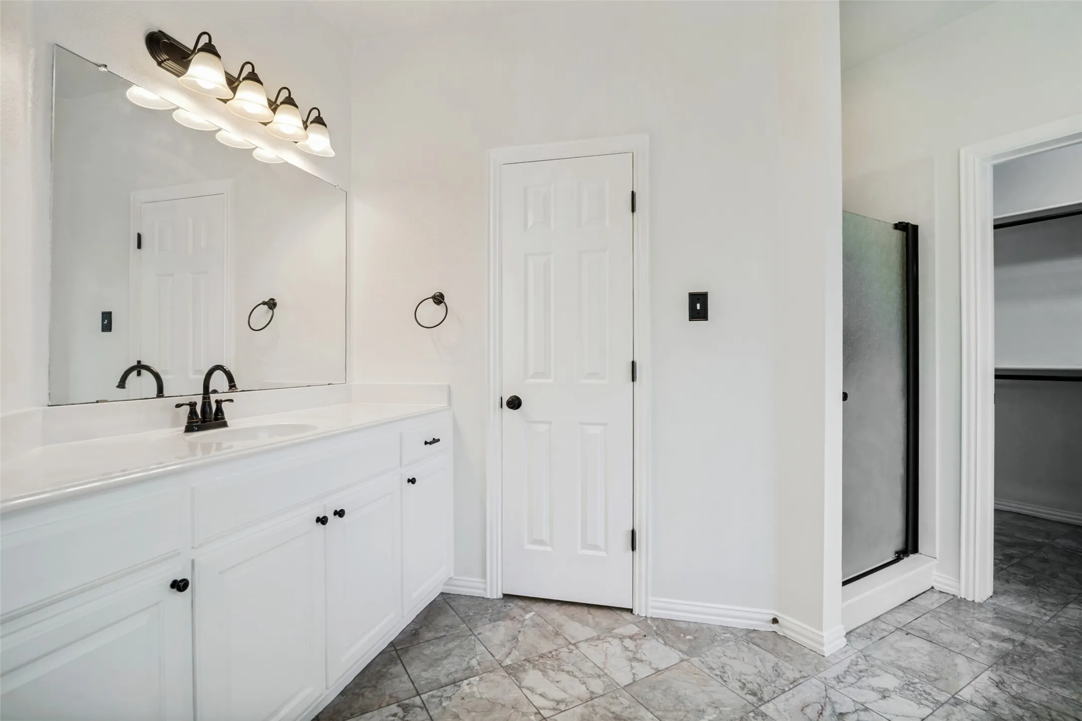 White vanity with an integrated sink and black hardware, large mirror with a five-light fixture, tiled flooring, and a glass-enclosed shower
