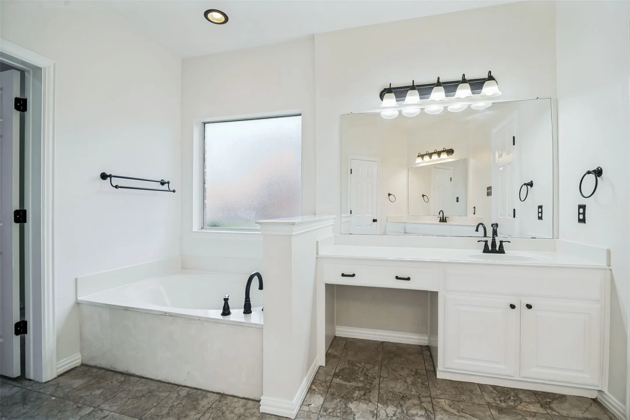 Spa-inspired primary bathroom featuring a freestanding soaking tub with integrated surround, dual-sink vanity with white cabinetry and brushed nickel hardware, large framed mirror with integrated lighting, and slate-finish flooring throughout