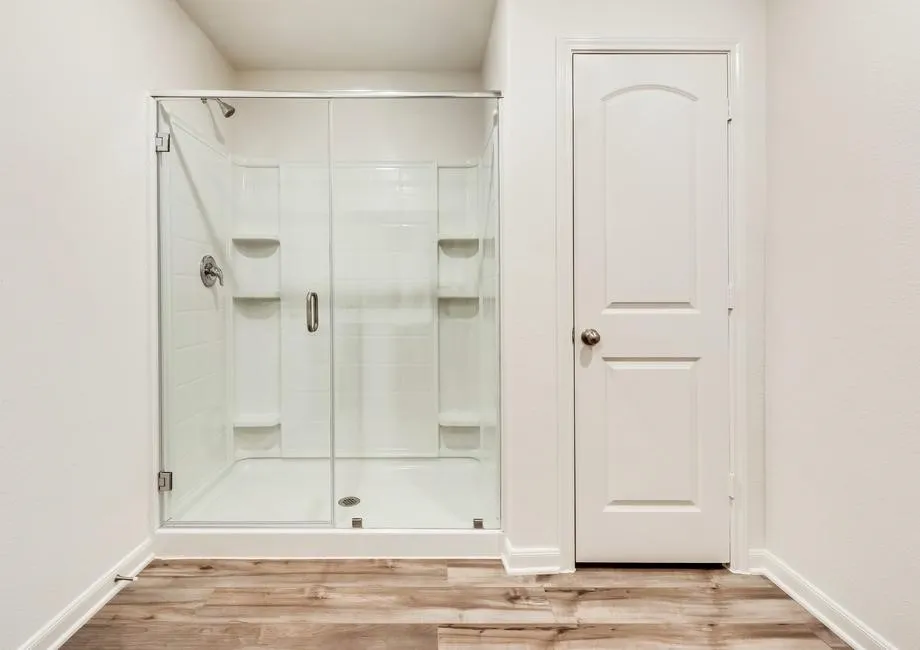 Enclosed shower with clear glass door, chrome fixtures, integrated shelving, and wood-finish flooring