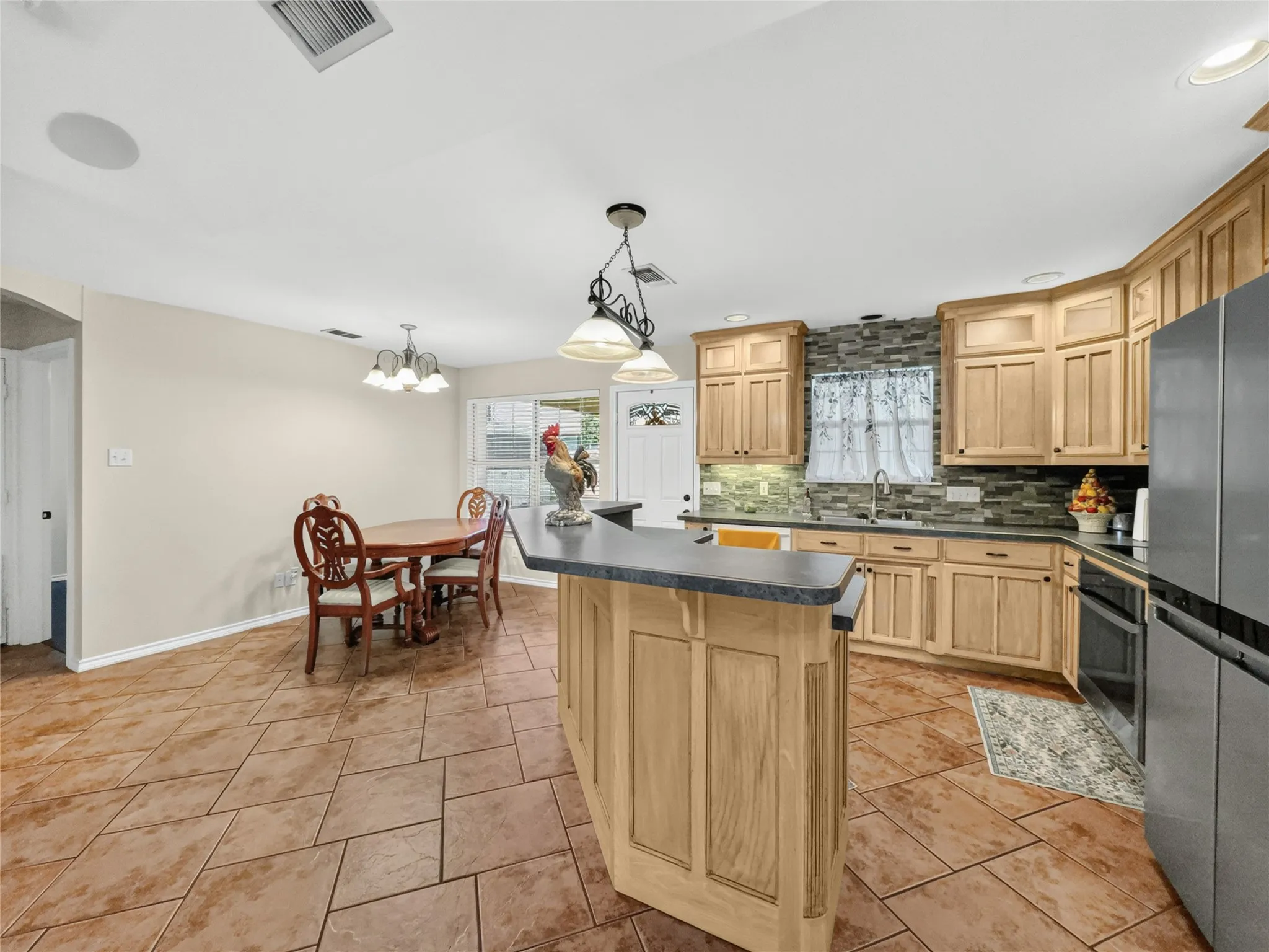 Kitchen featuring natural wood cabinetry, stone-look tile flooring, a multi-tone tile backsplash, built-in stainless steel appliances, and an island with counter seating