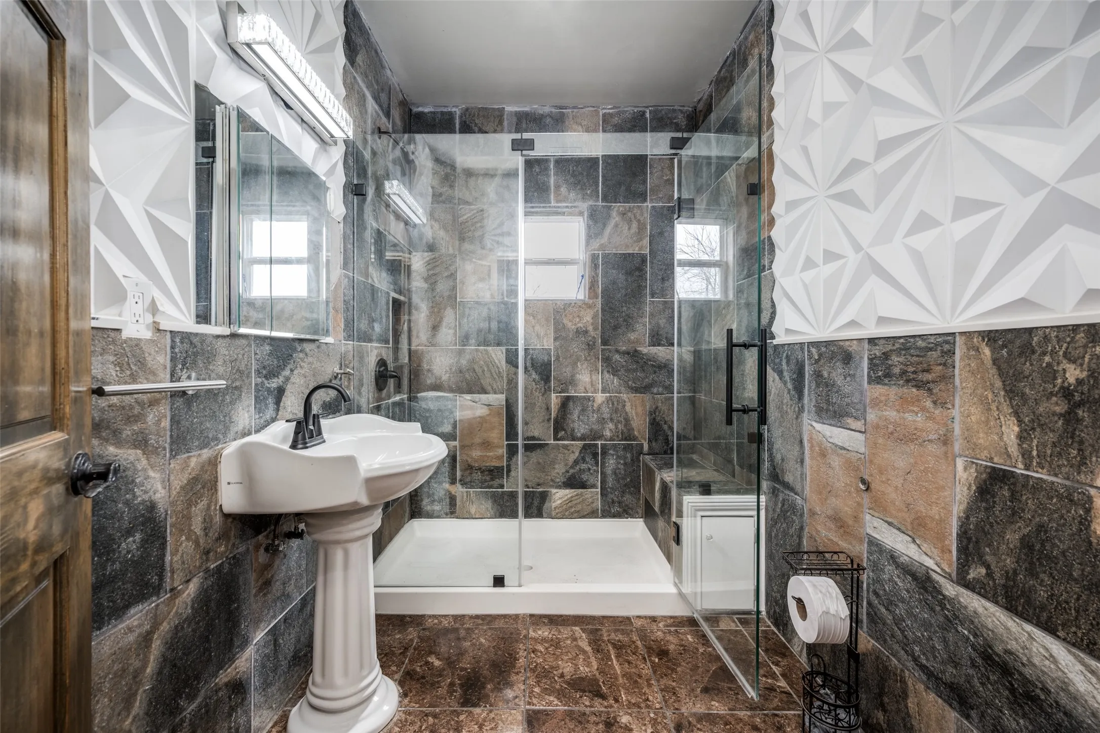 Pedestal sink with dark-toned fixtures, floor-to-ceiling natural stone wall tiling, and a glass-enclosed shower with a built-in bench