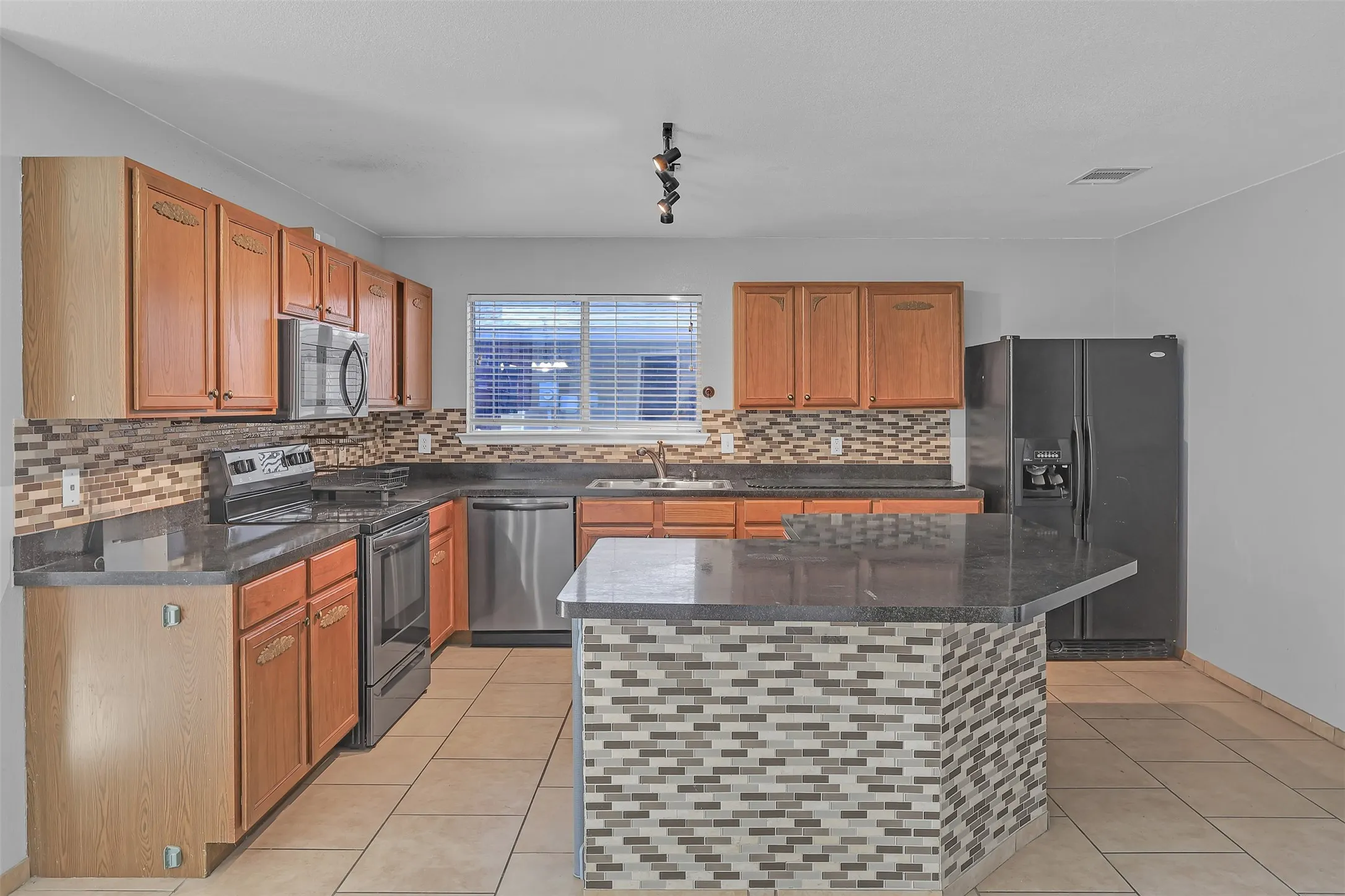Kitchen with appliances with stainless steel finishes, brown cabinets, light tile patterned flooring, dark stone counters, and tasteful backsplash