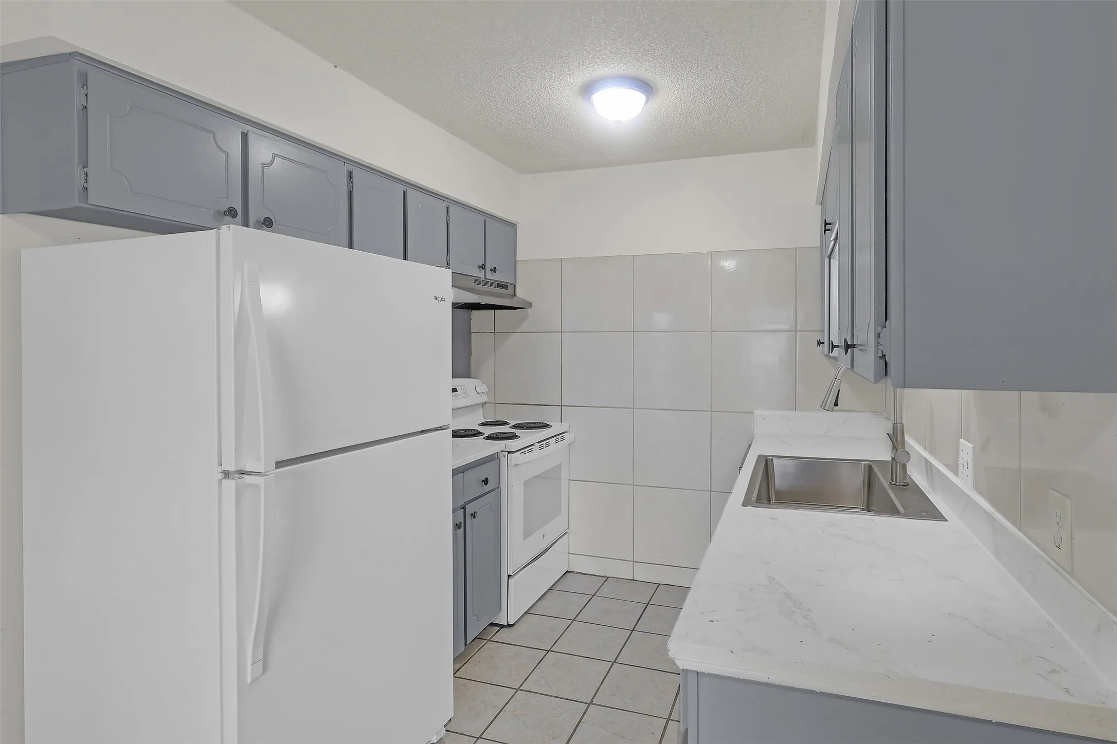 Kitchen featuring white appliances, gray cabinetry, under cabinet range hood, a textured ceiling, and light tile patterned floors