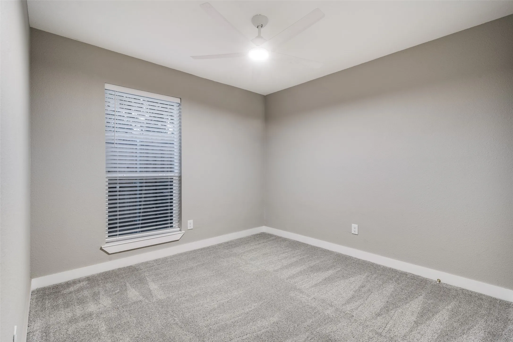 Neutral gray wall paint, white trim, gray carpet flooring, white window blinds, and a white ceiling fan with integrated lighting