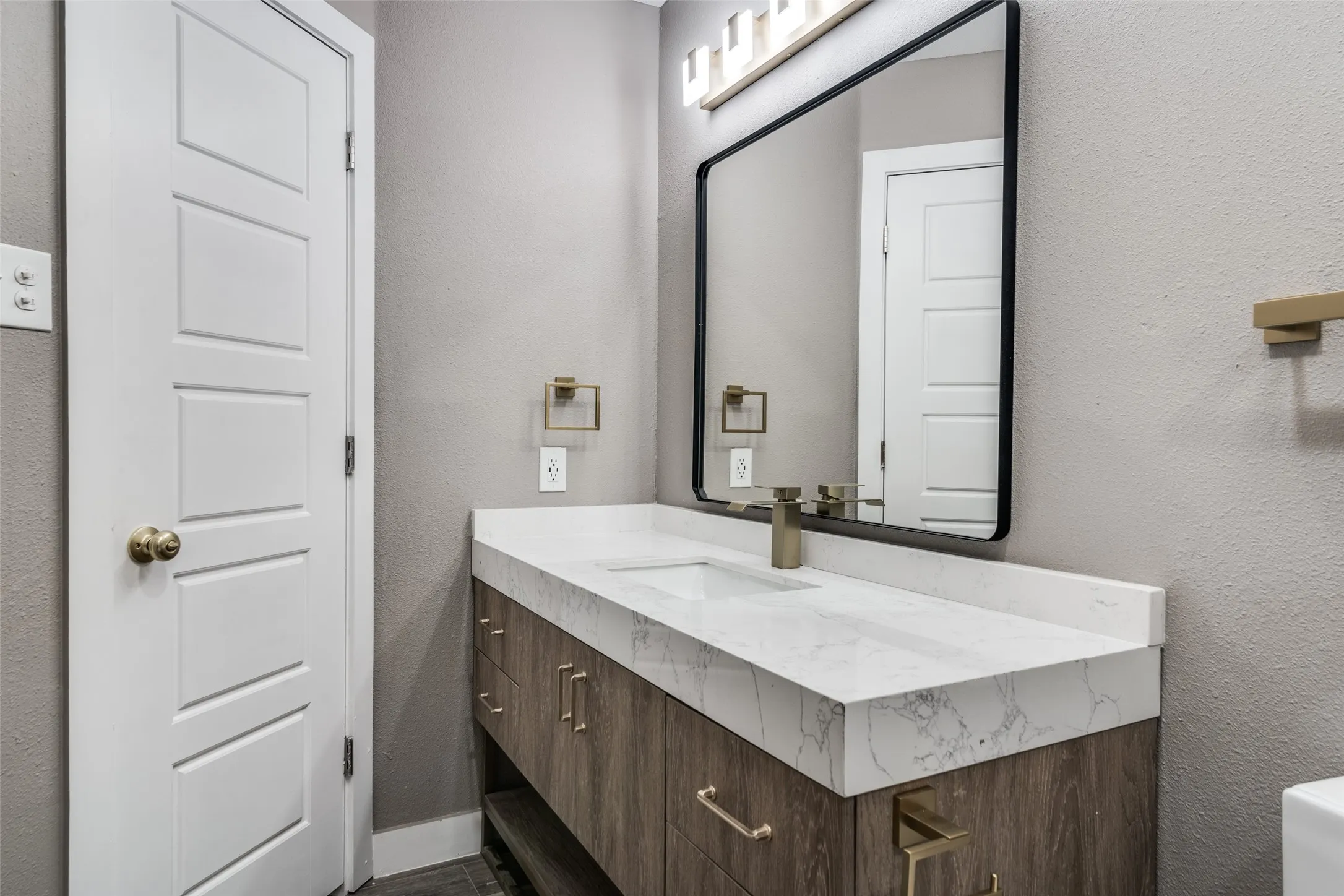 Floating wood-finish vanity with gold-tone hardware, white countertop with integrated sink, rectangular mirror with a black frame, and a gold-tone faucet