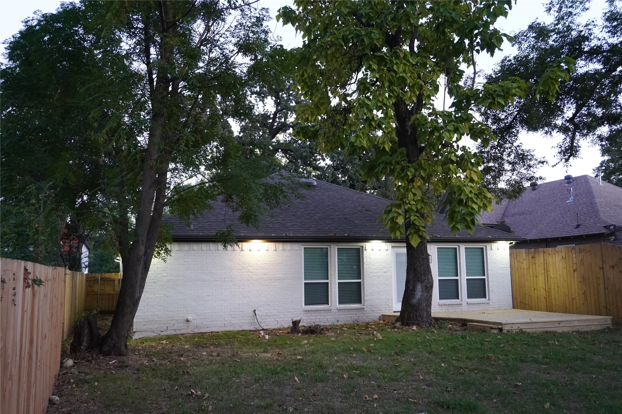 Rear exterior featuring a wood fence, multiple trees, a dark shingle roof, a white brick facade, and a built-in wood deck