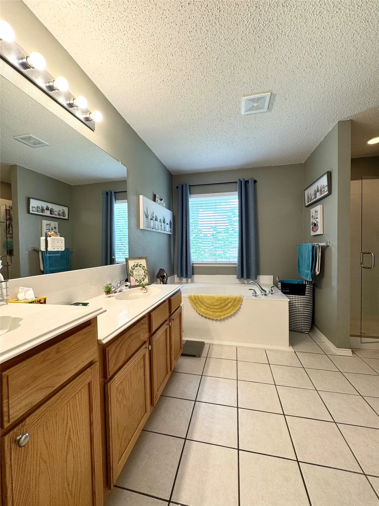 Dual sink vanity with light wood-finish cabinetry, built-in soaking tub, tiled flooring, and an enclosed shower with a glass door
