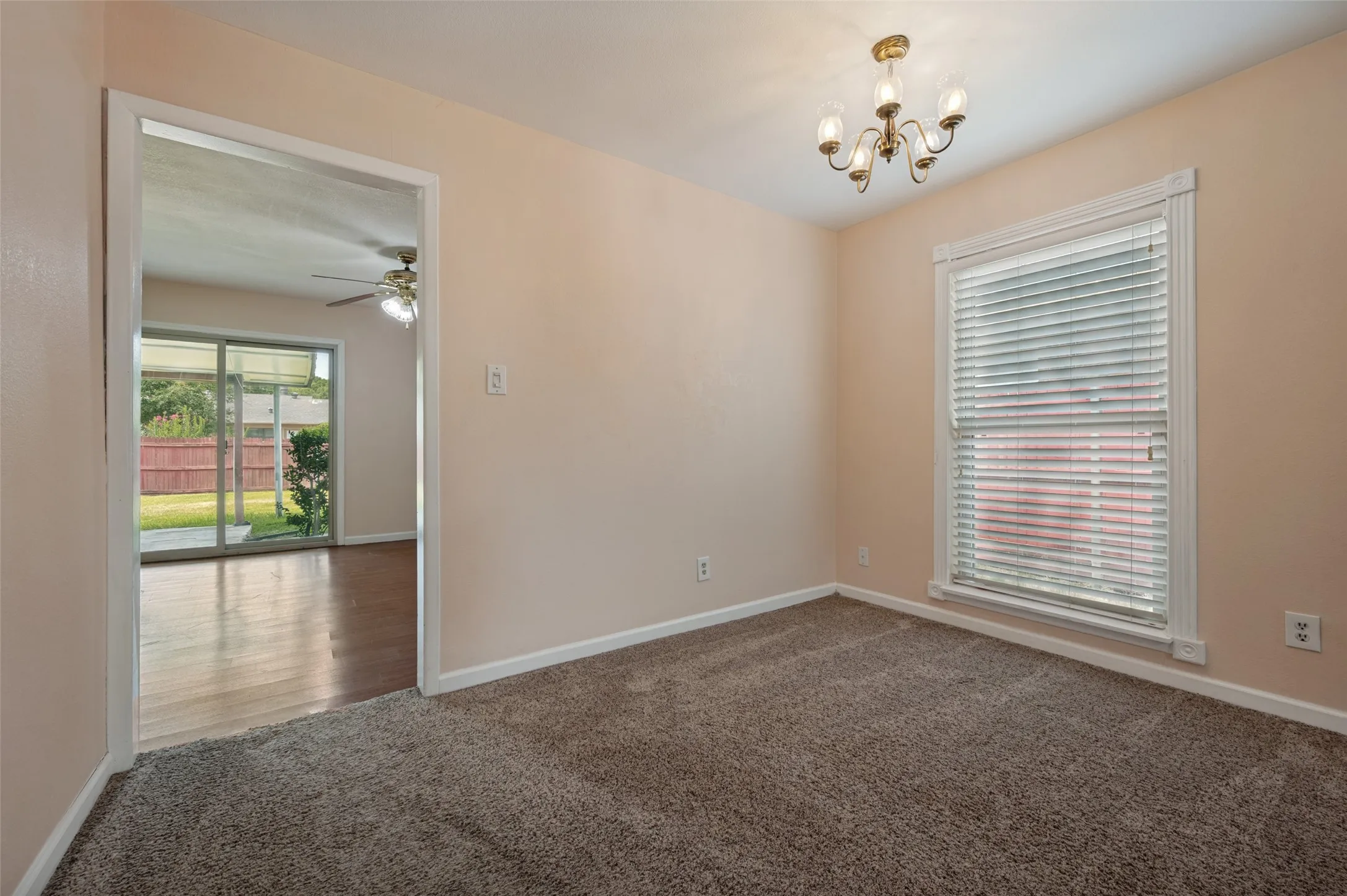 Carpeted room with a brass chandelier, window with blinds, and a doorway leading to a room with wood-finish flooring and sliding glass doors