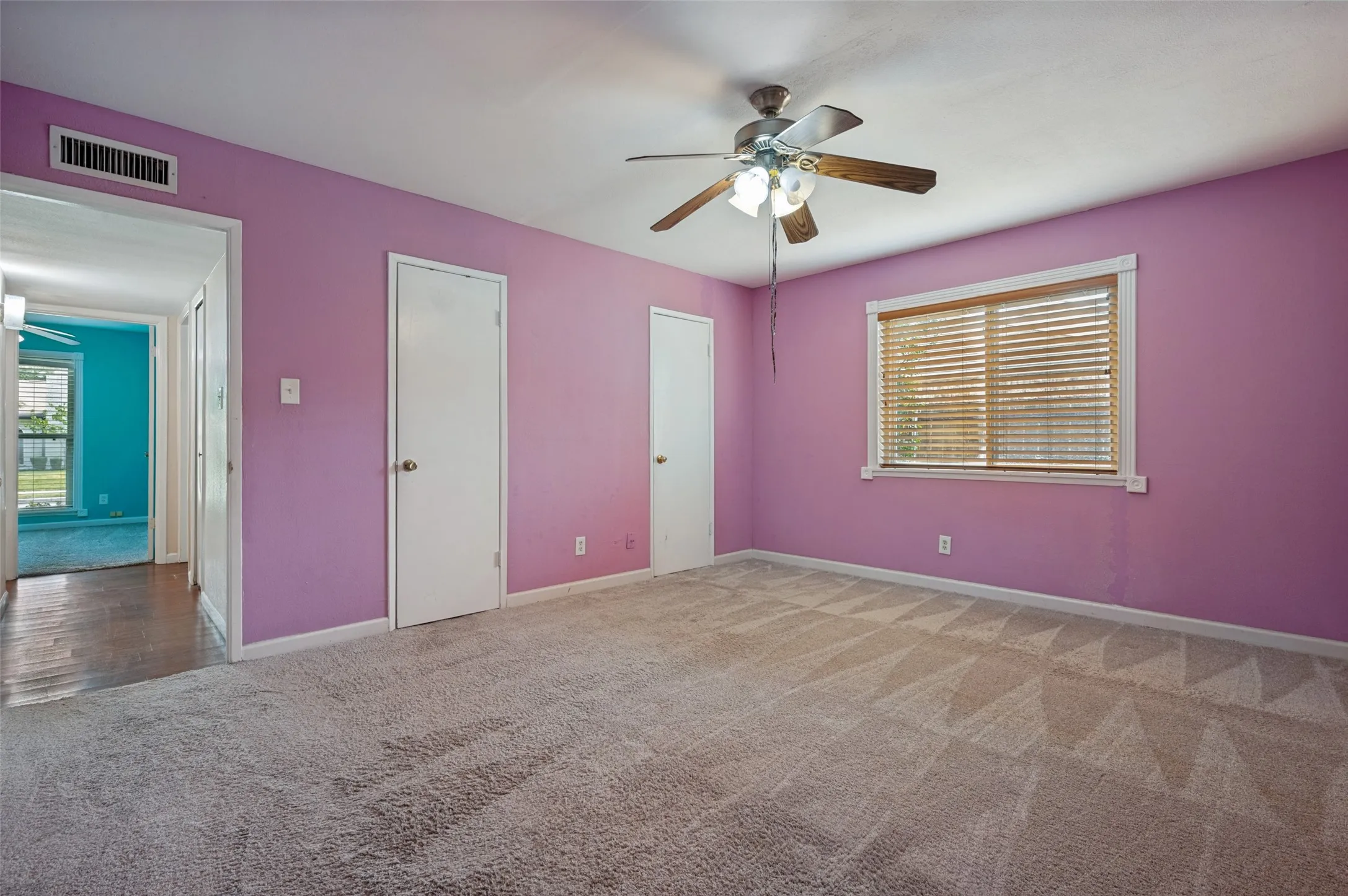Spacious room with plush carpeting, a ceiling fan with integrated lighting, two white panel doors, and a window with horizontal blinds