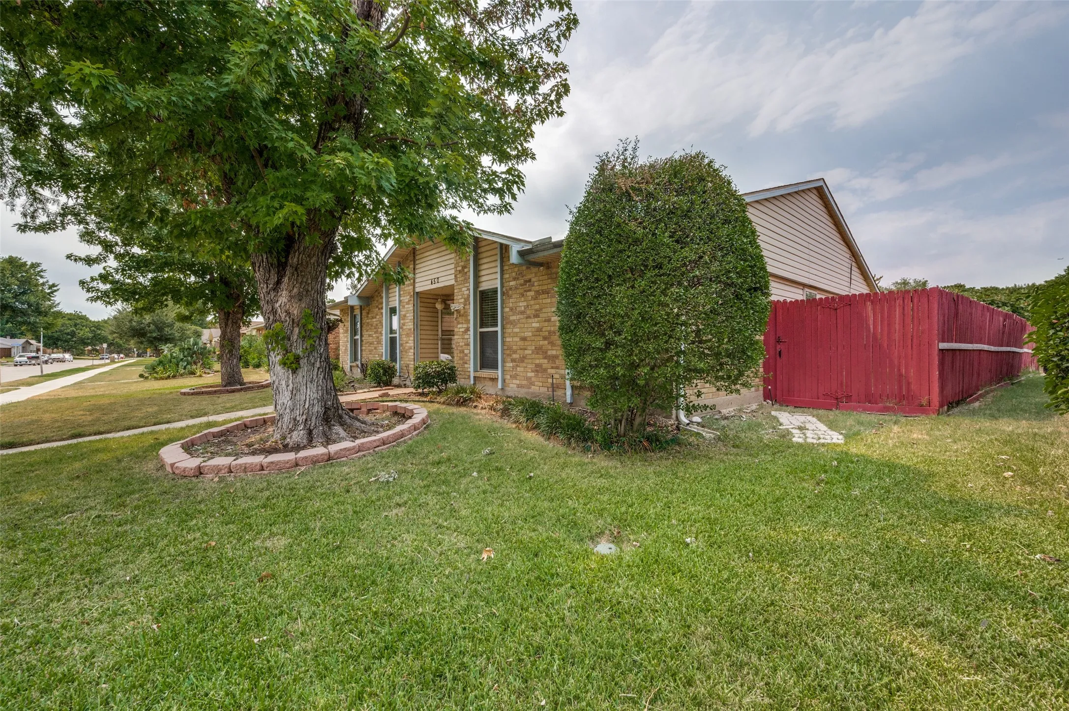 Brick exterior with horizontal siding accents, mature shade trees with decorative brick edging, red privacy fencing, and a manicured lawn