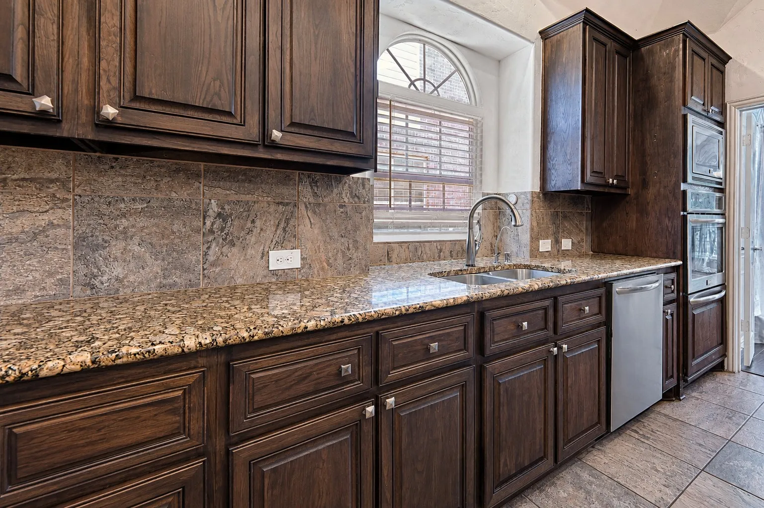 Granite countertops, dual basin stainless steel sink with gooseneck faucet, dark wood cabinetry, tile backsplash, and stainless steel built-in double ovens
