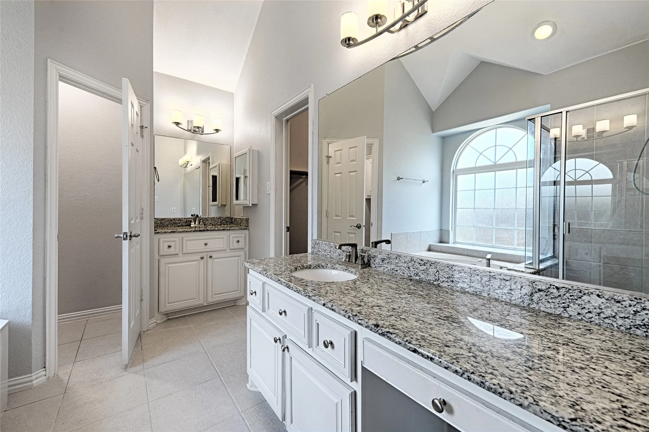 Dual vanities with granite countertops, tile flooring, an arched window, a built-in bathtub, and a glass-enclosed shower