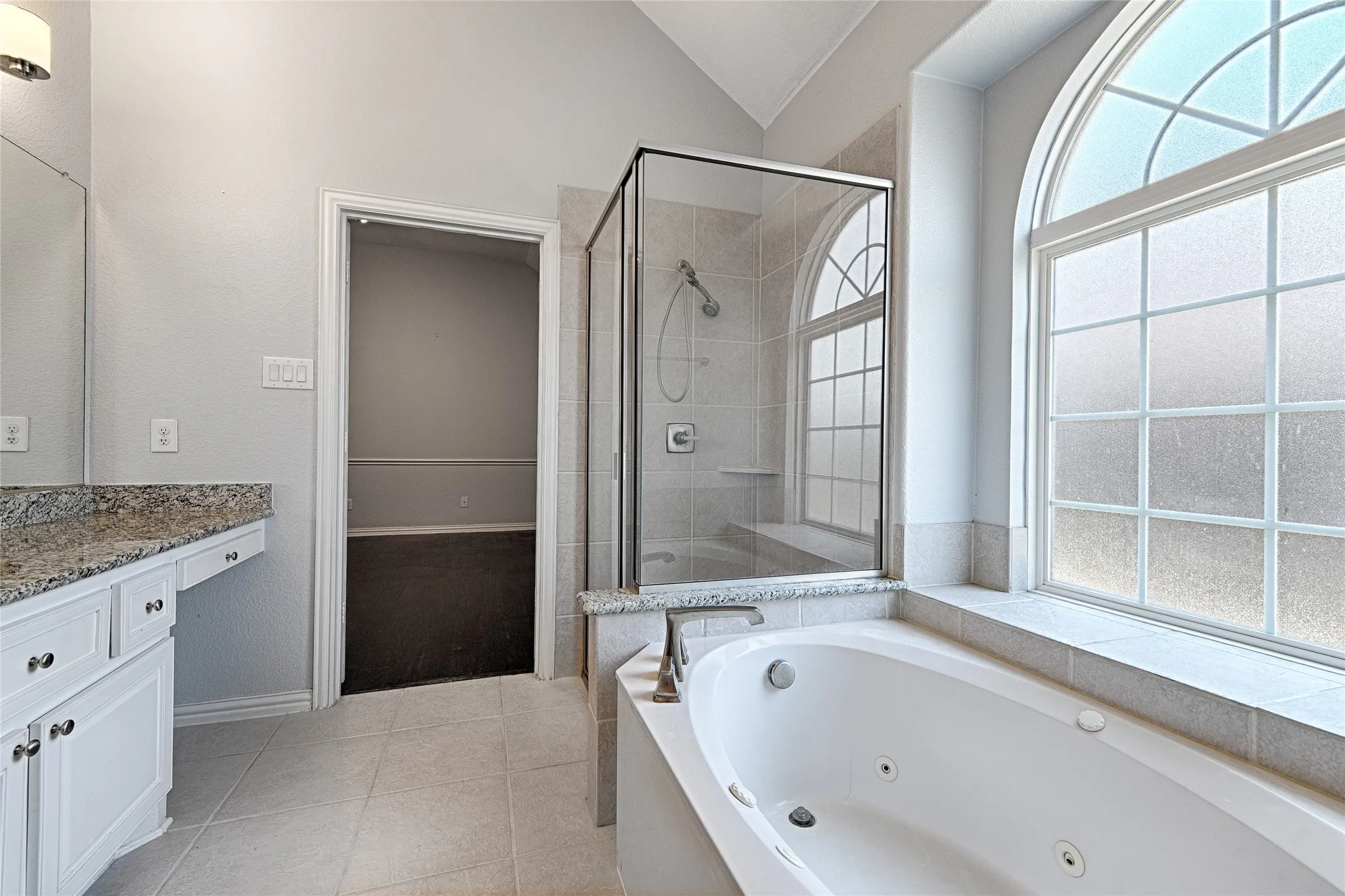 Spacious bathroom with a walk-in shower, jetted bathtub, dual-pane arched window, and vanity with stone countertop