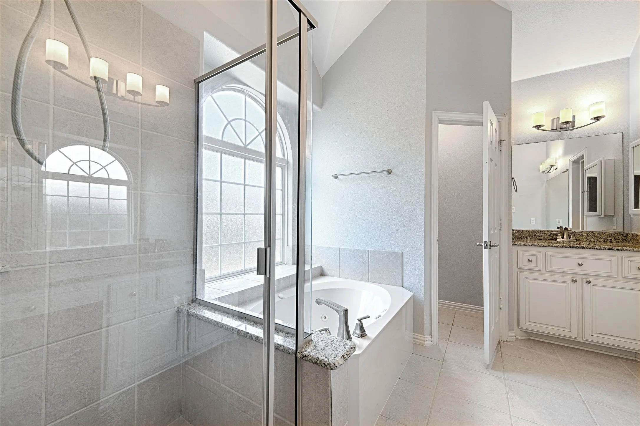 Bathroom featuring a built-in tub, separate glass-enclosed shower, arched window, vanity with granite countertop, and tiled flooring