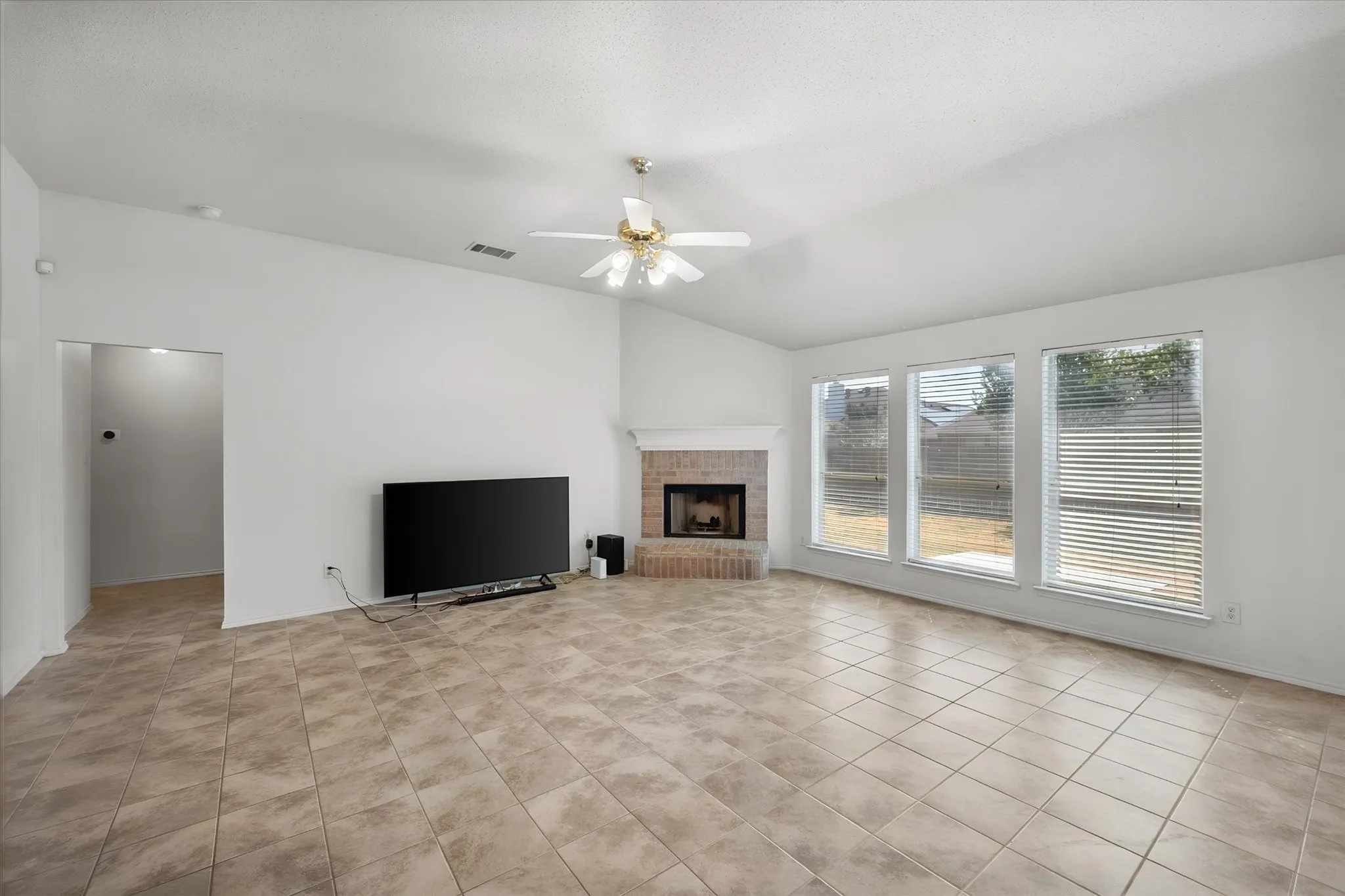 Spacious living area featuring vaulted ceilings, a ceiling fan, tiled flooring, a brick-front fireplace, and three large windows
